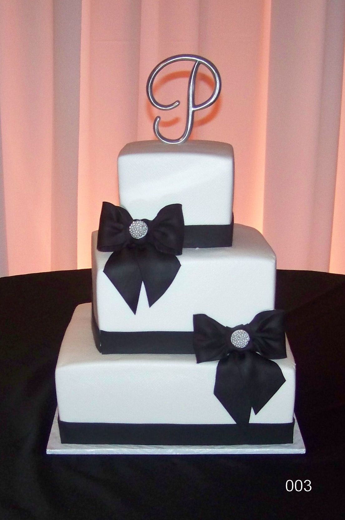 Three-tier white cake with black bows and a silver topper against a pink backdrop