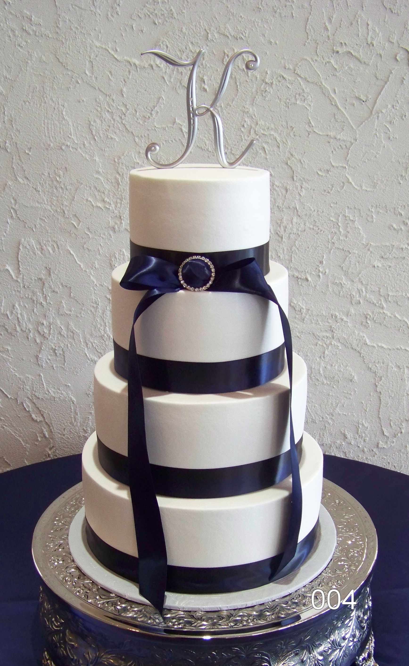 Three-tier white cake with dark blue ribbon and silver monogram topper on a decorated silver stand