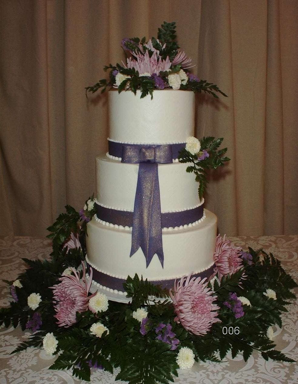 Three-tier white wedding cake with purple ribbons and pink flowers on a floral table display