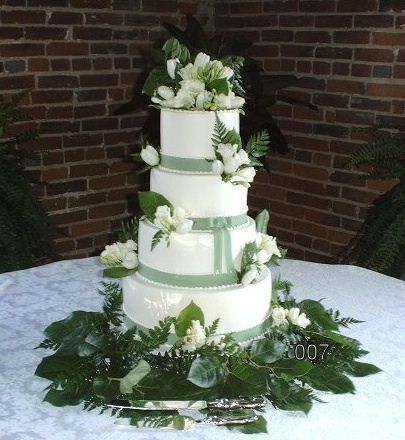 Four-tier white wedding cake with green ribbons and white flowers on a table against a brick wall