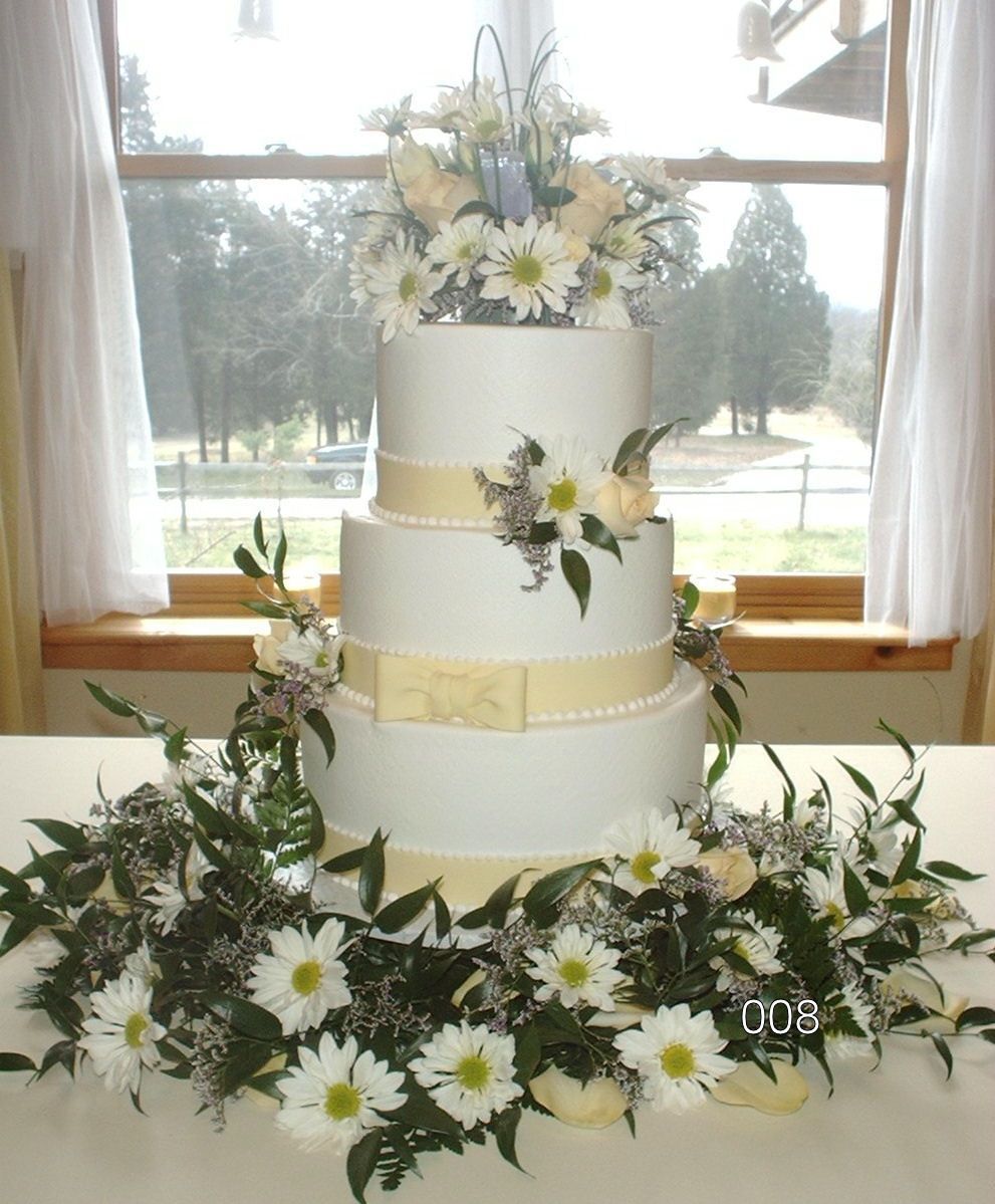 Three-tier white wedding cake with cream ribbons and white daisy floral decorations by a window