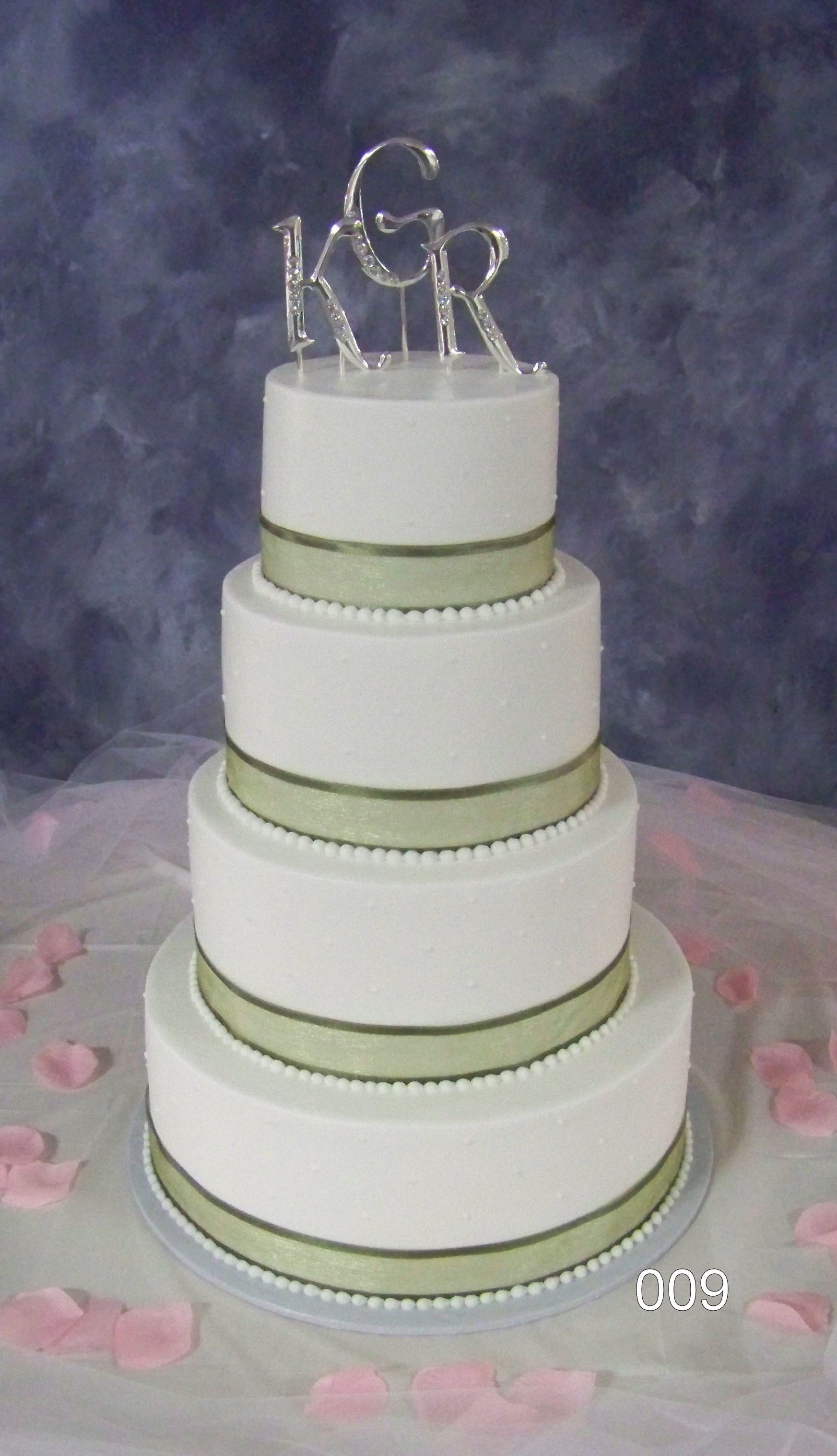 Four-tier white wedding cake with green ribbon trim and a silver monogram topper on a pink tablecloth