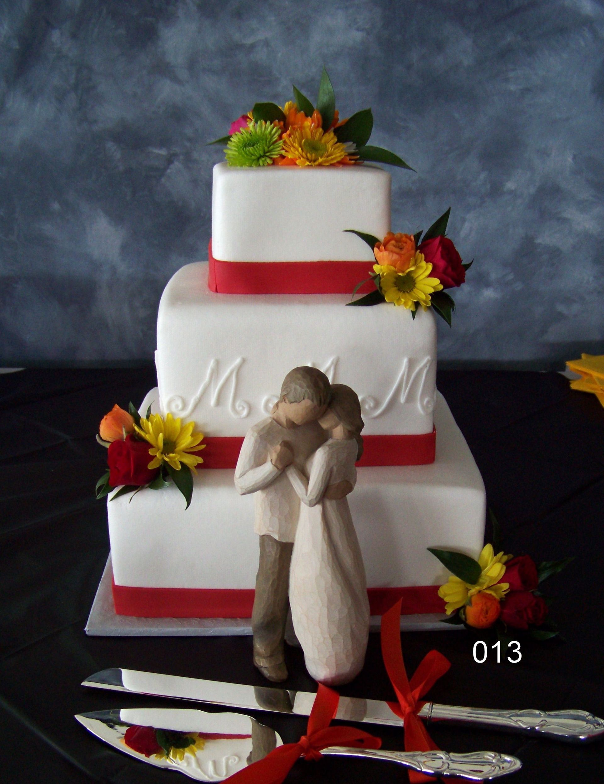 Three-tier white cake with red ribbon trim and orange floral toppers, plus a small angel figurine in front.