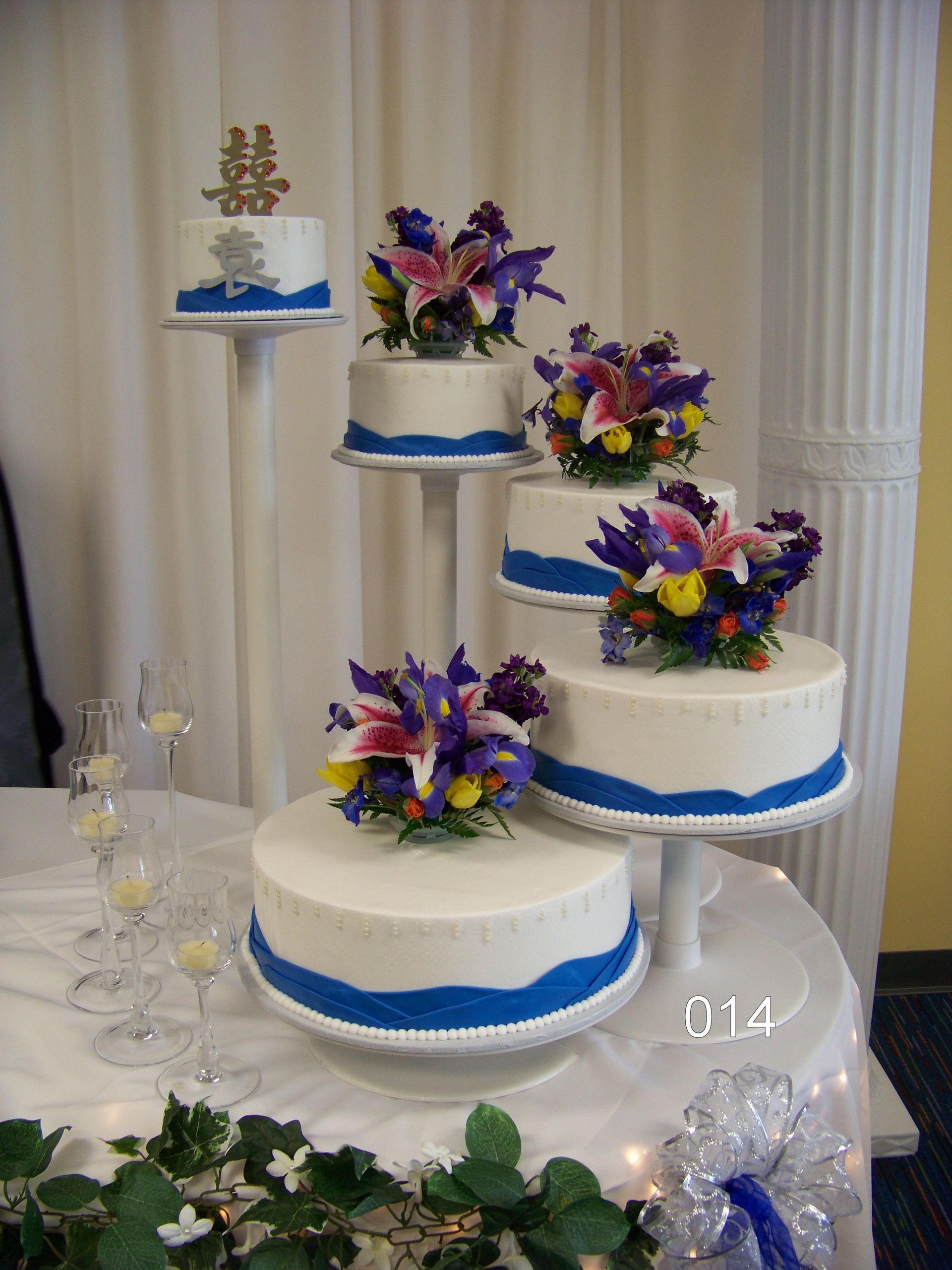 Four-tier white wedding cake with purple flowers and blue ribbon trim on a decorated table
