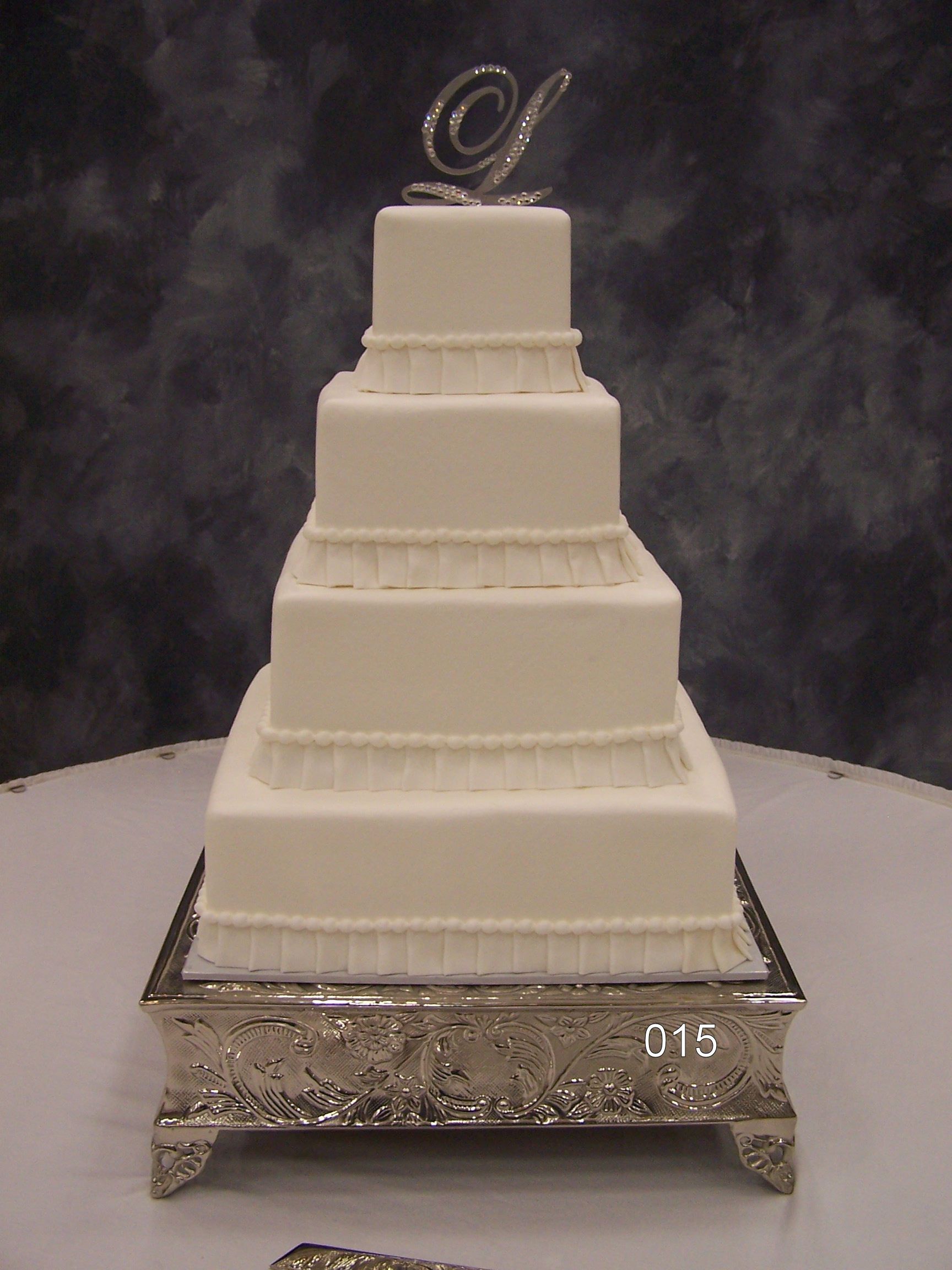 White tiered wedding cake on an ornate silver stand, topped with a decorative monogram.