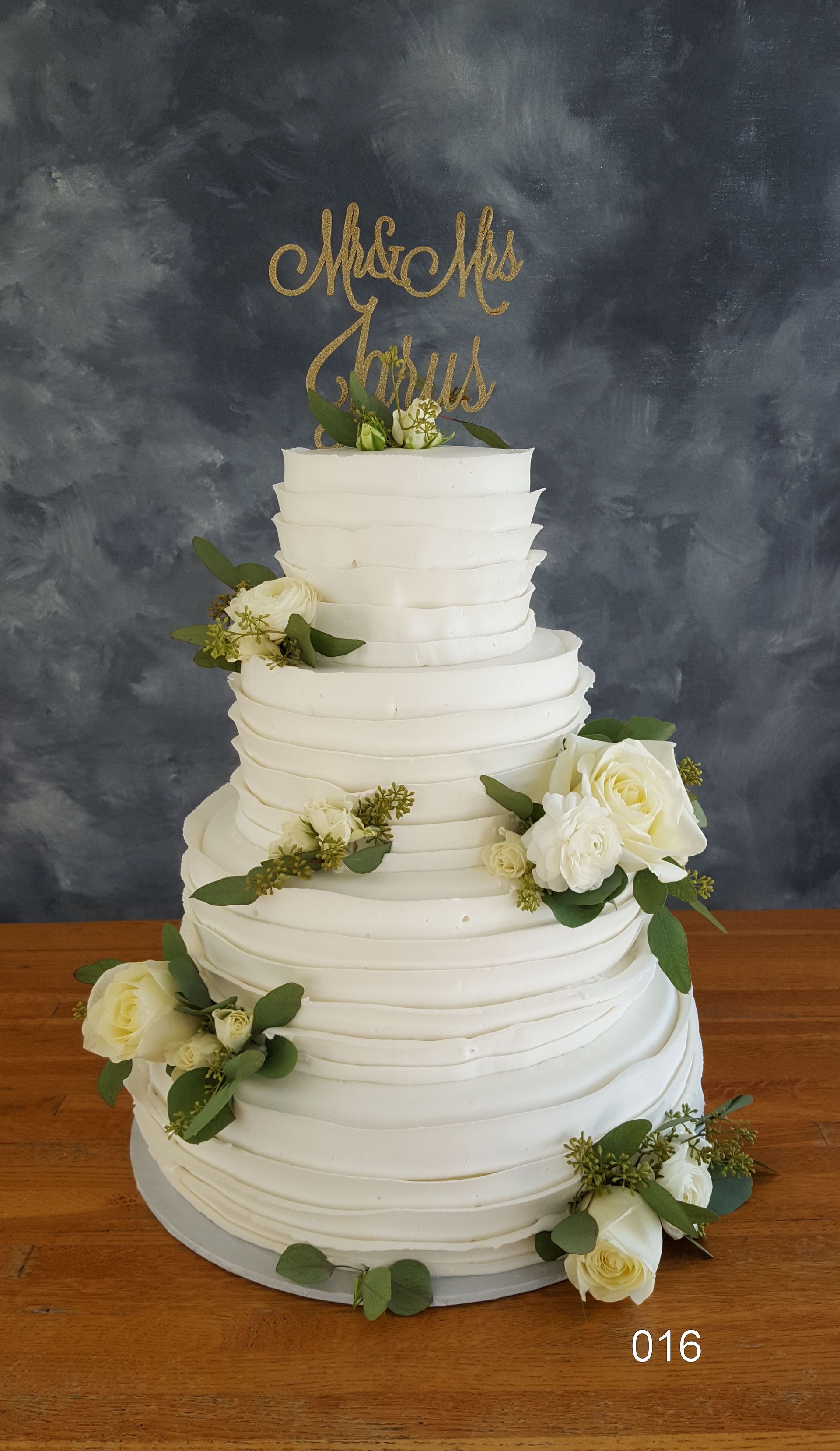 Three-tier white wedding cake with white roses and greenery, topped with a gold “Mrs & Mrs” topper.
