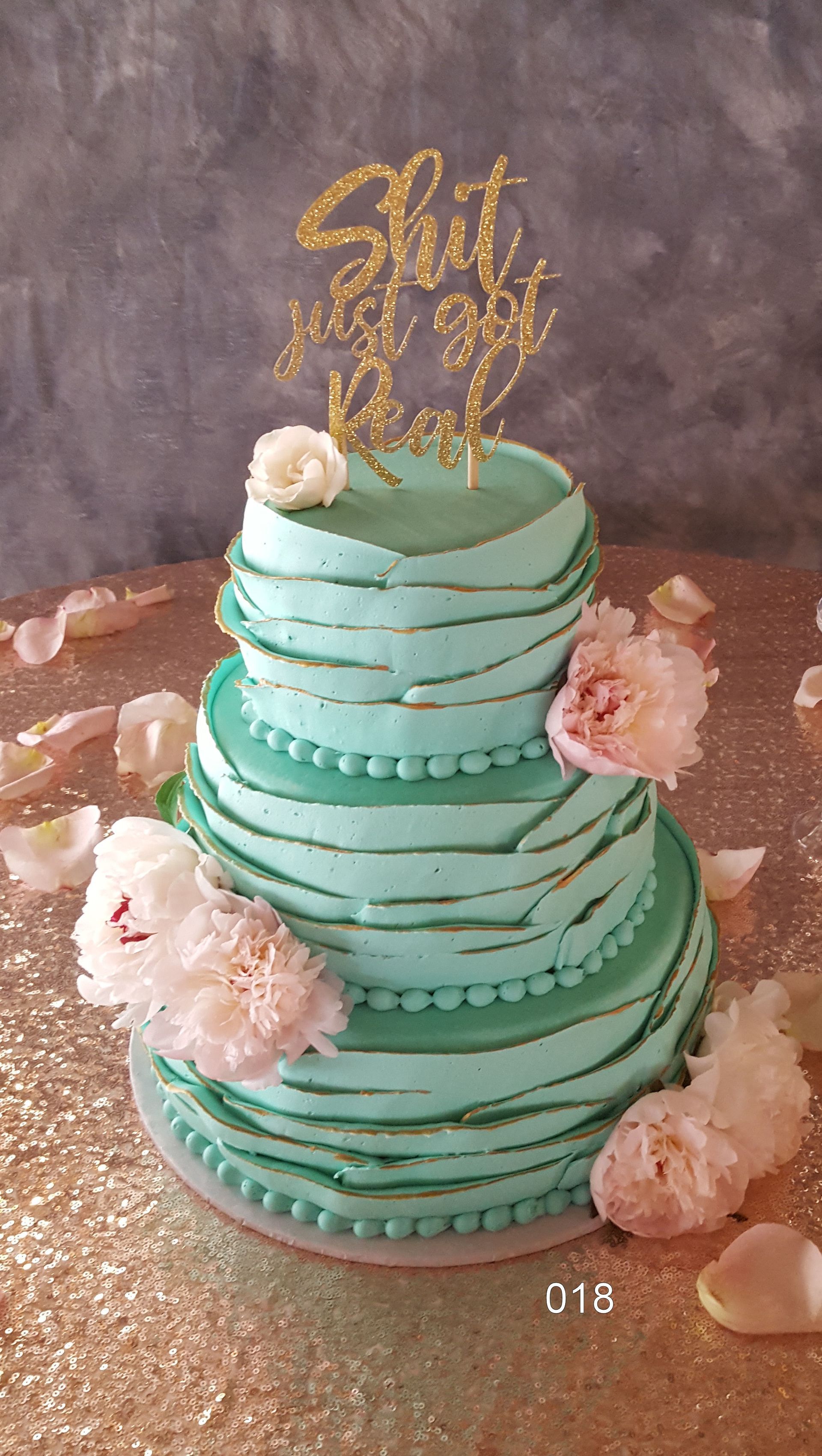 Mint-green tiered cake with pink flowers and gold topper on a glittery rose-gold table