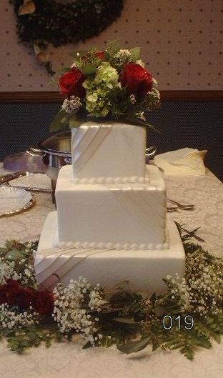 Three-tier white wedding cake with red and white flowers, surrounded by greenery on a decorated table.