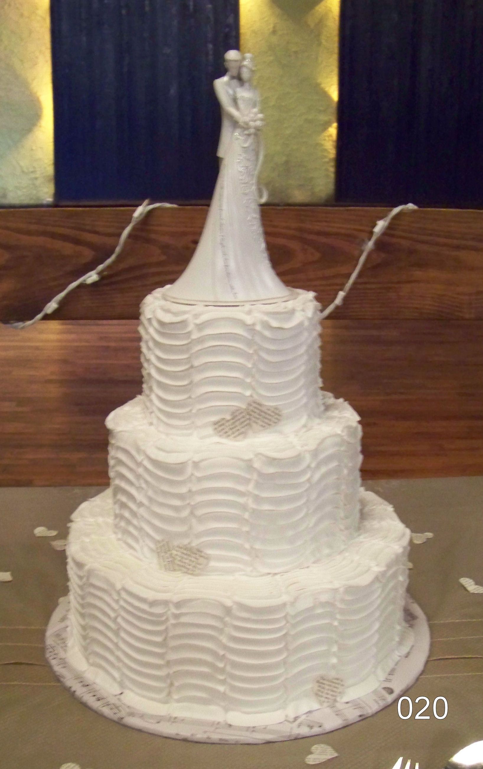 White tiered cake topped with a small figurine, displayed on a wooden table with golden backdrop lights.