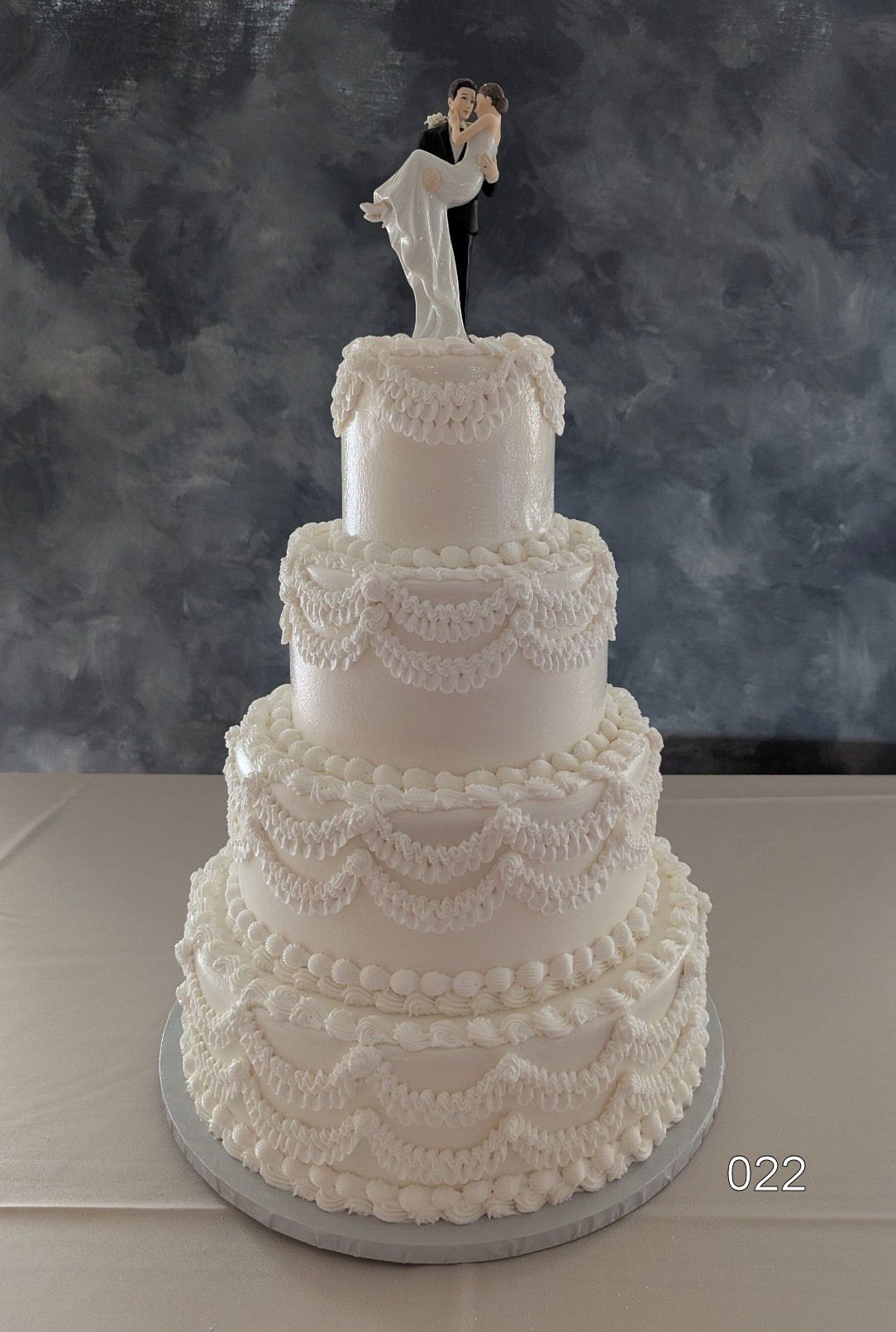 Four-tier white wedding cake with lace-like frosting and a bride topper on a gray backdrop