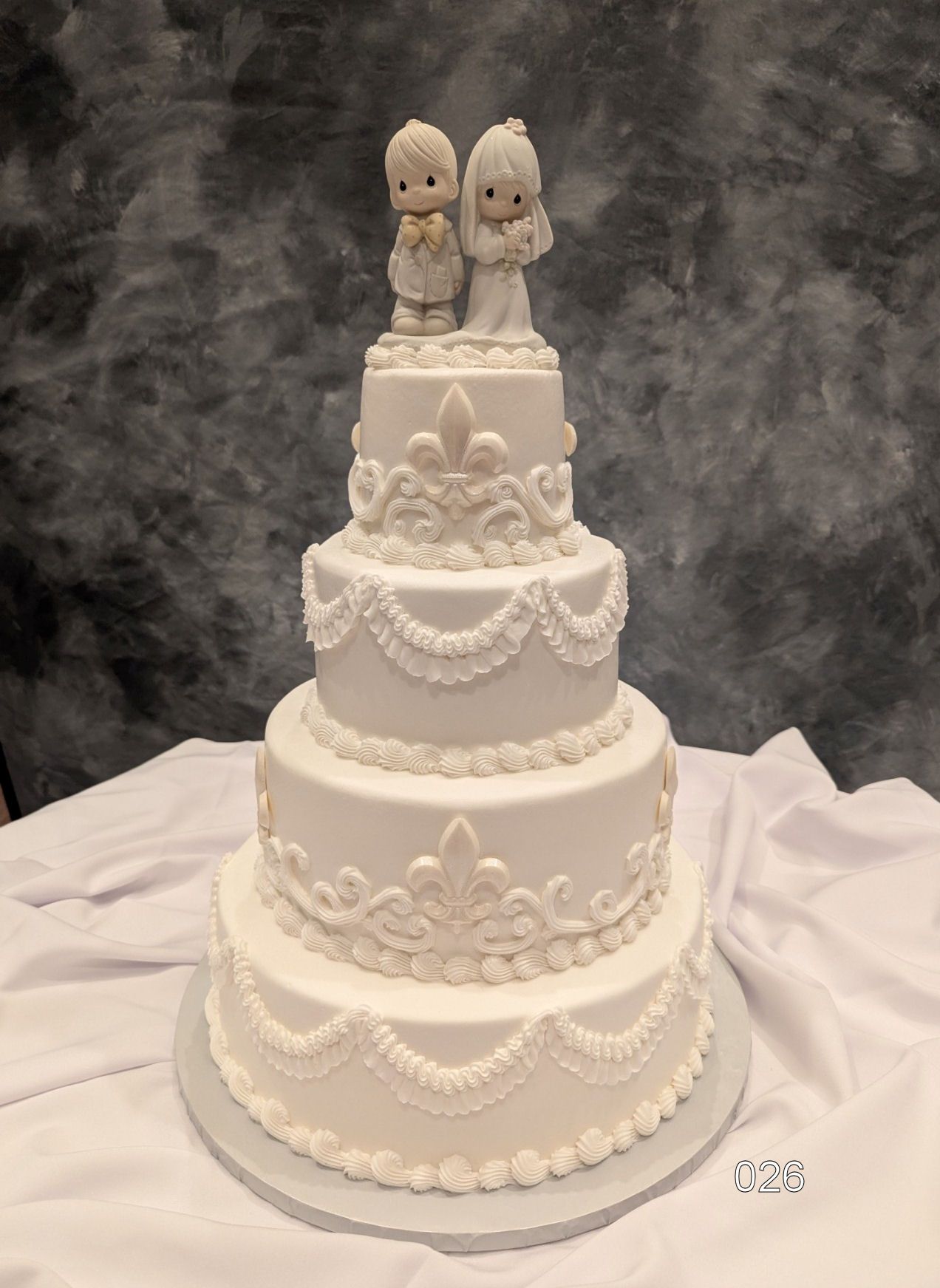 White tiered wedding cake with ornate piping and two small figurines on top, against a gray backdrop