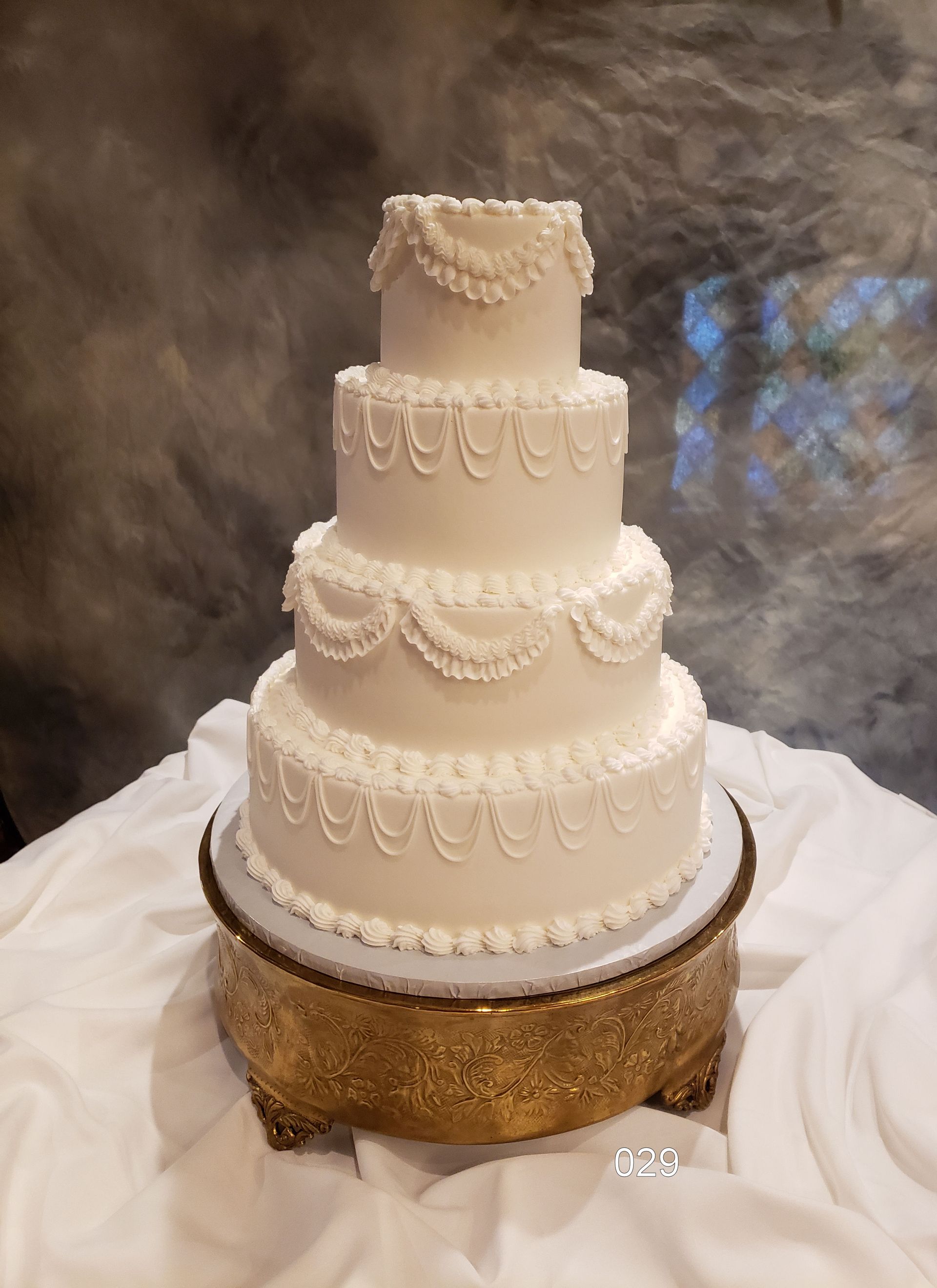 Four-tier white wedding cake with lace piping on a gold stand, set on draped white fabric