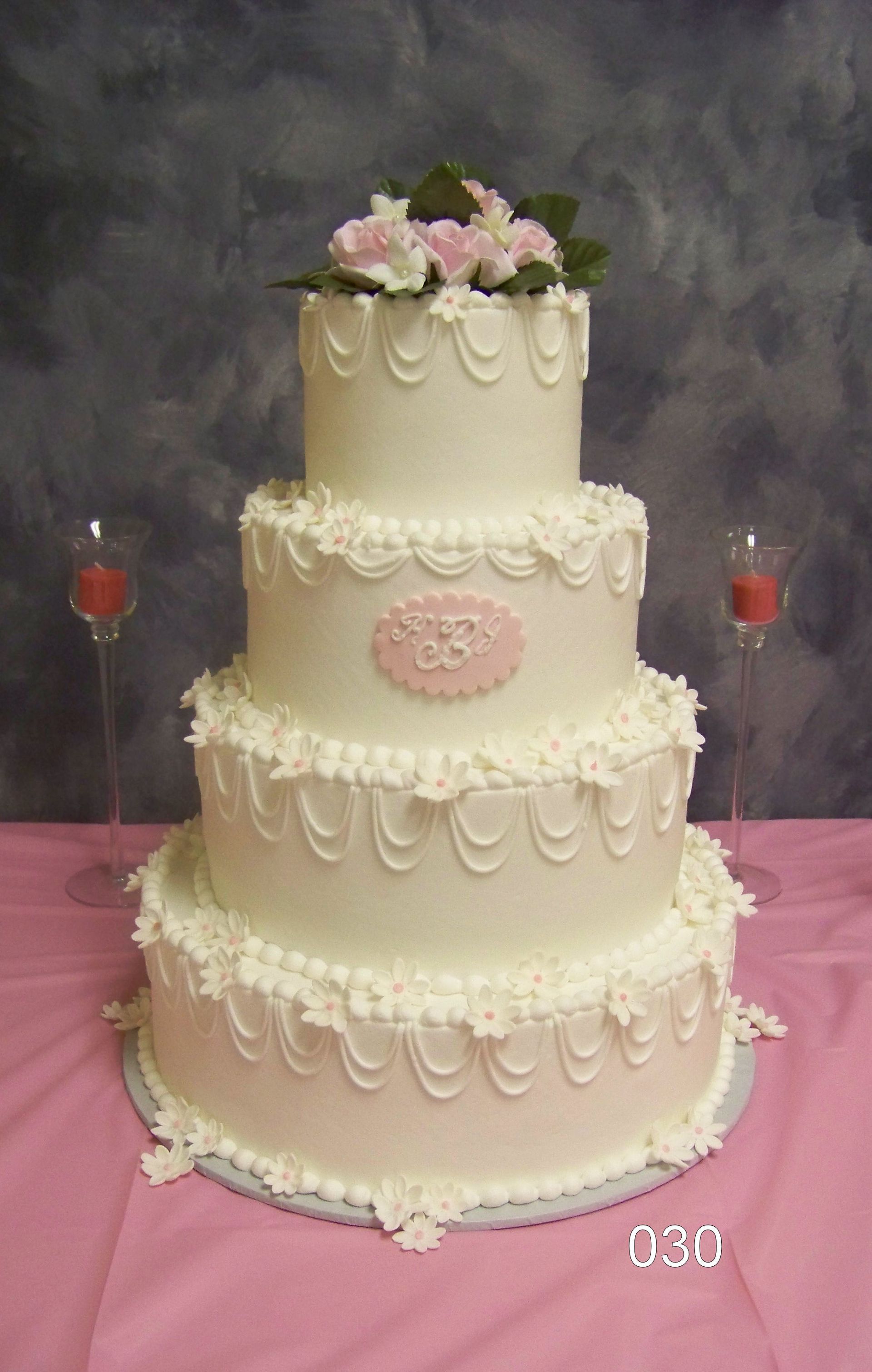 Three-tier white wedding cake with pink flowers on top and ornate icing details, on a table with red candles