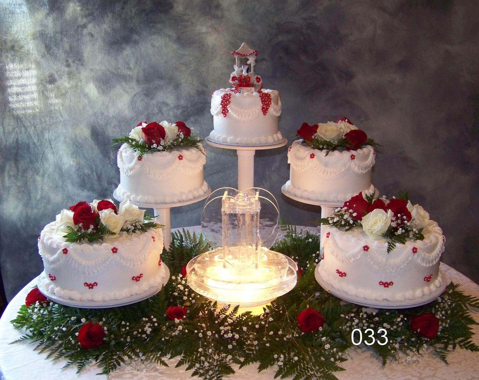 Five white cakes with red roses arranged around a lit centerpiece on a decorative table