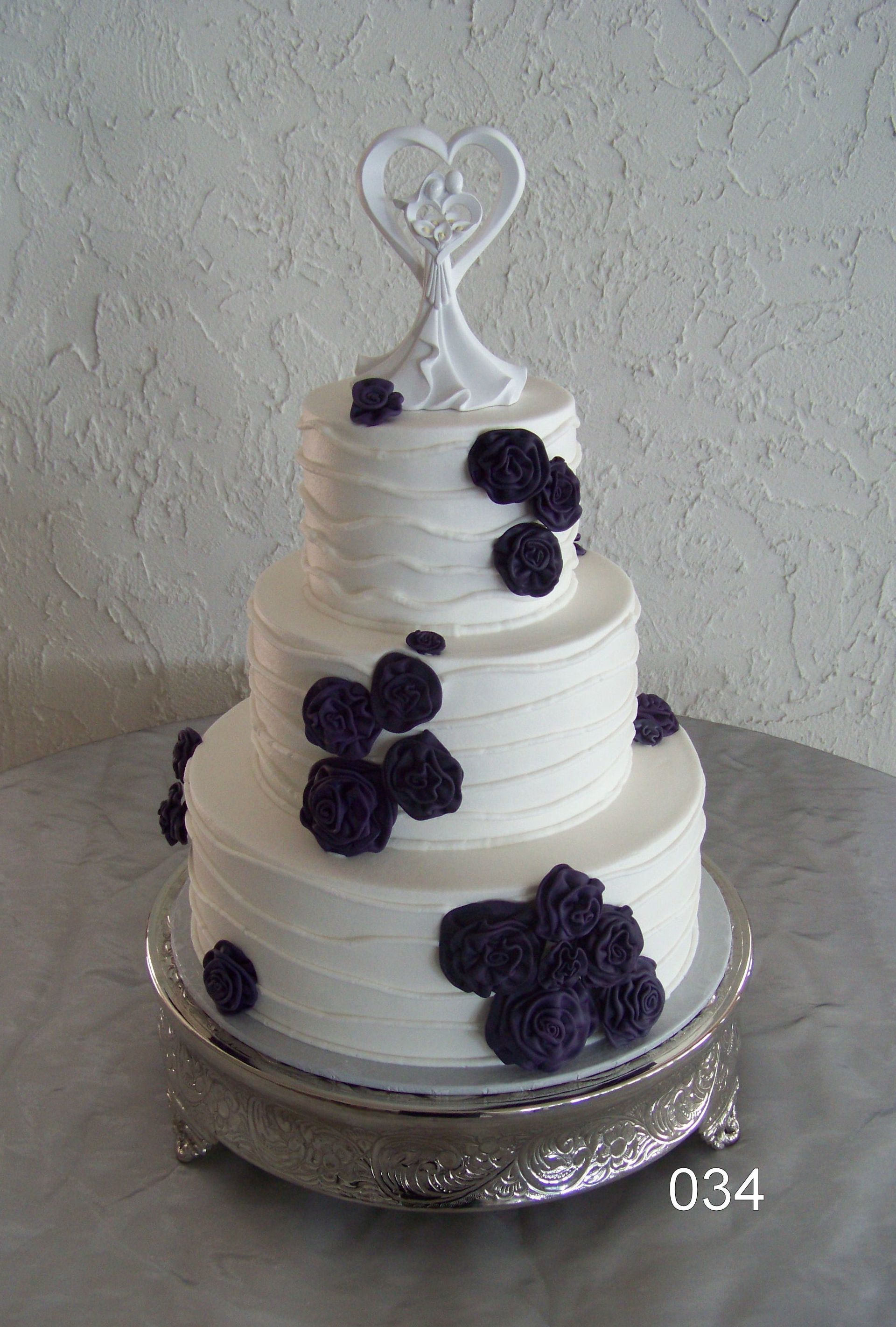 Three-tier white wedding cake with purple flowers and a heart topper on a silver stand