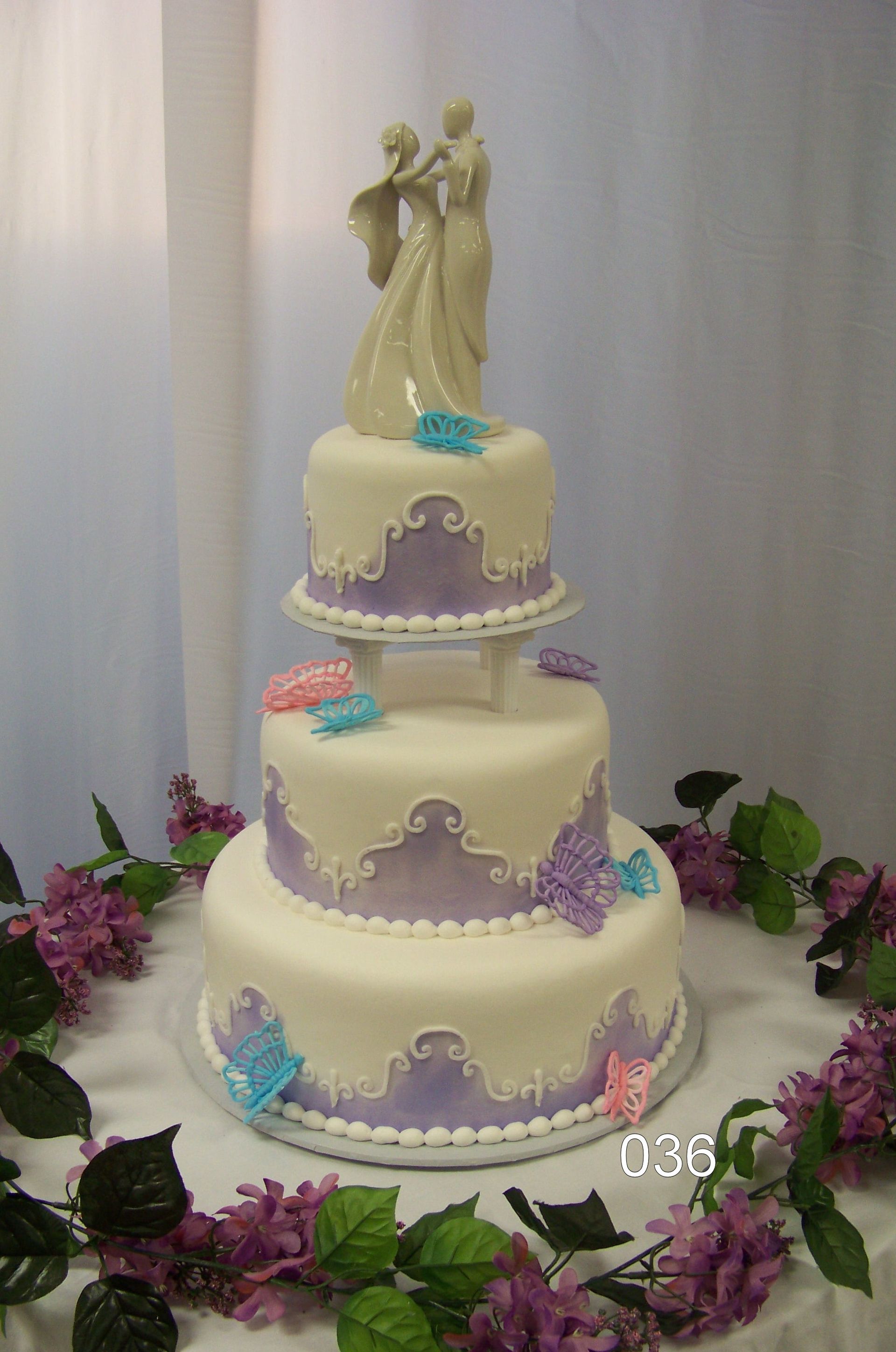 Three-tier white wedding cake with purple trim, blue flowers, and a bride and groom topper, surrounded by flowers