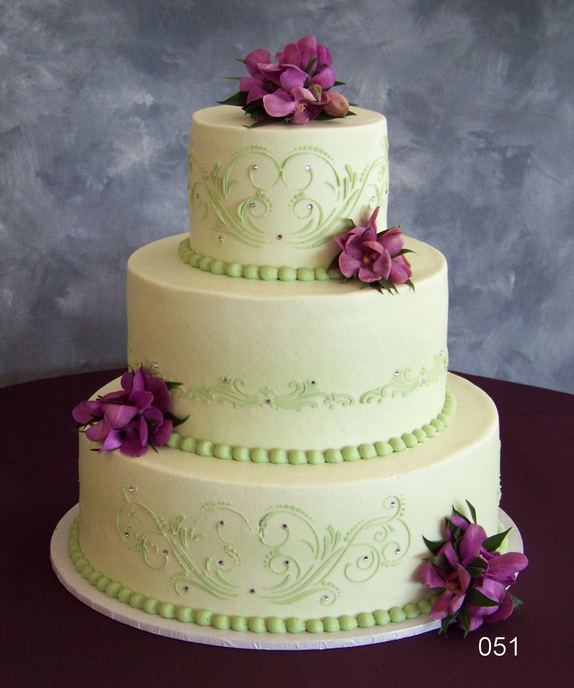 Three-tier white cake with green trim and purple flowers on a gray backdrop