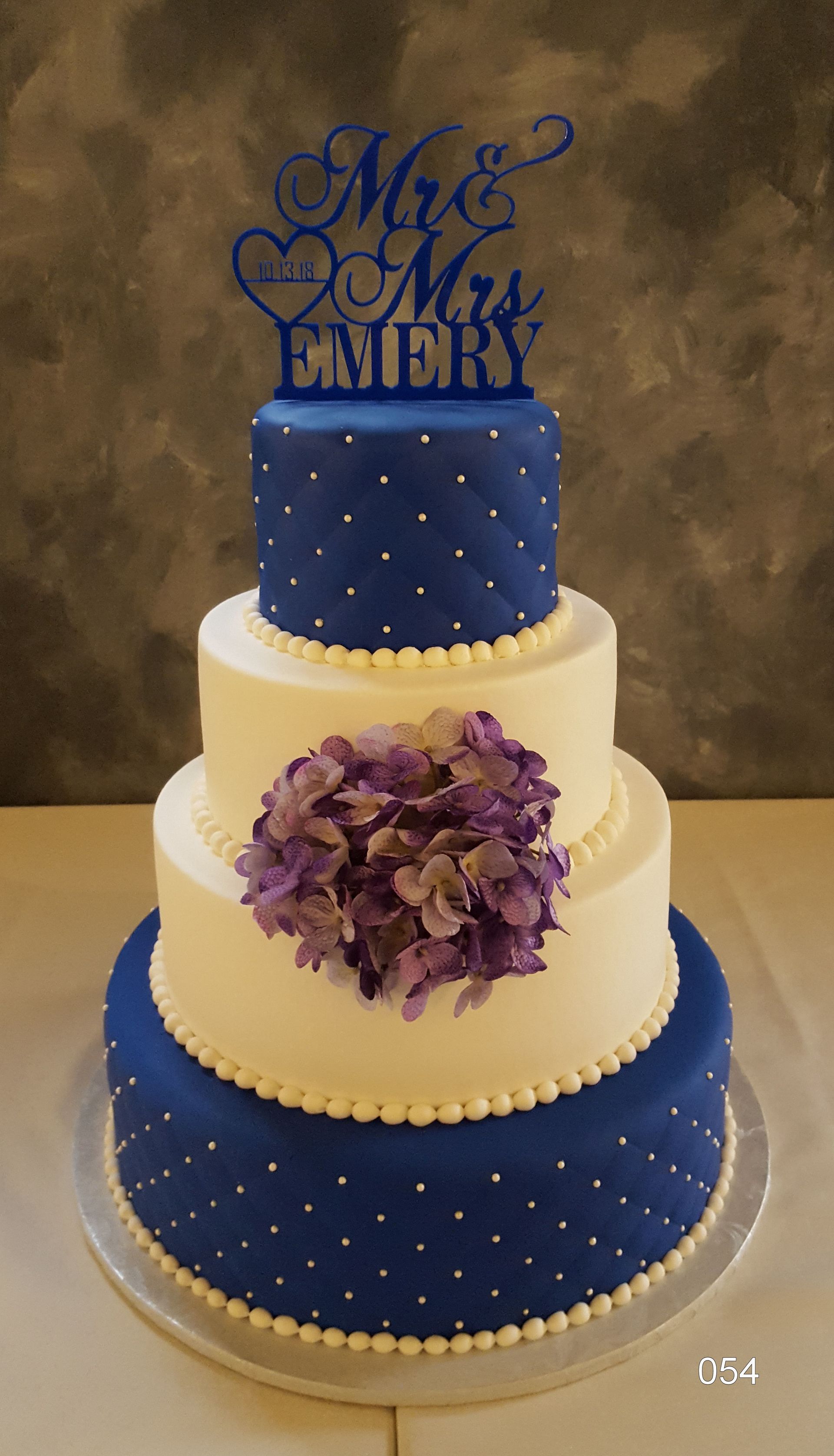 Three-tier blue and cream wedding cake with purple flowers and a blue topper reading “Mr & Mrs”