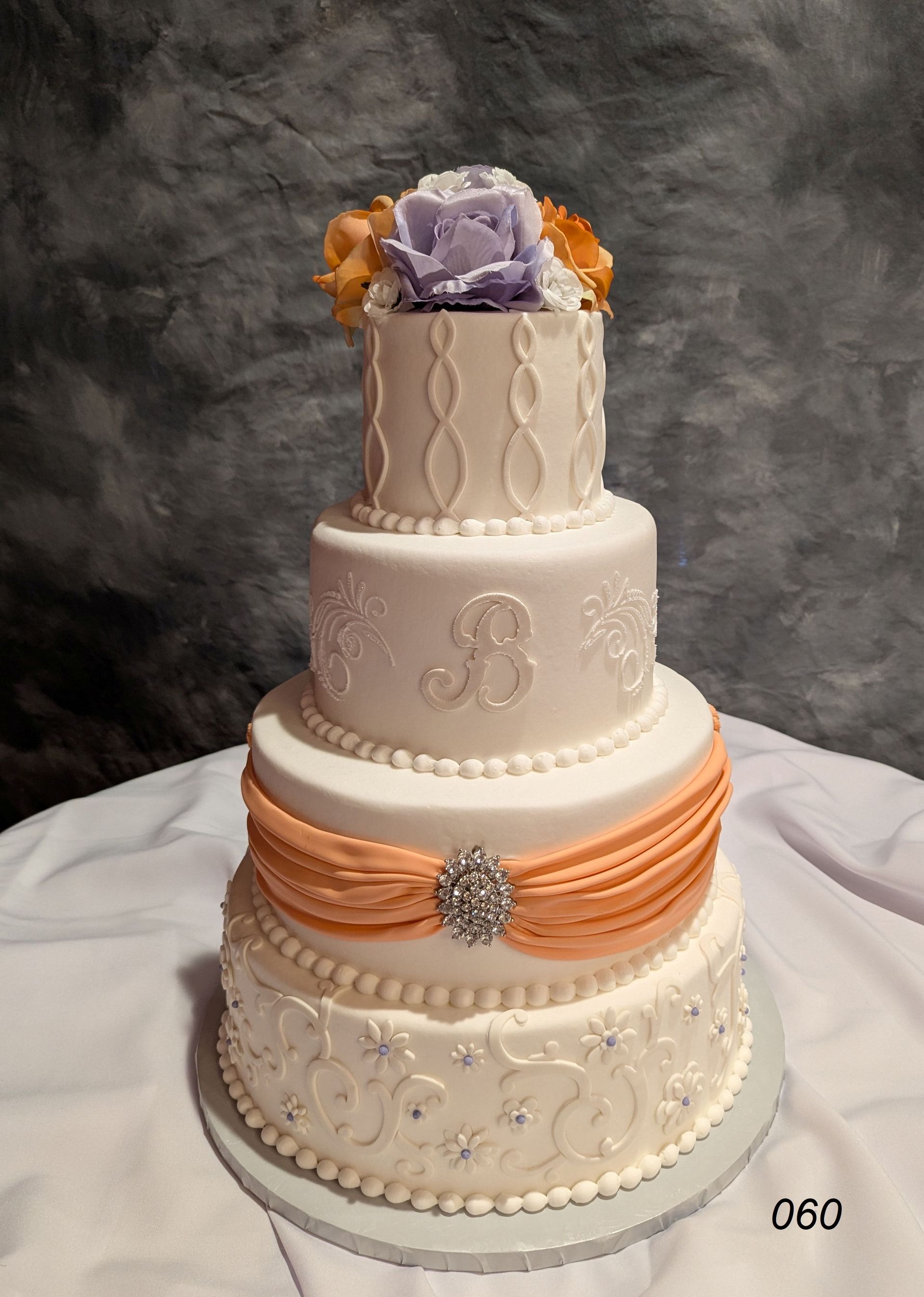 Four-tier white wedding cake with orange ribbon and silver accents, topped with a lavender flower arrangement
