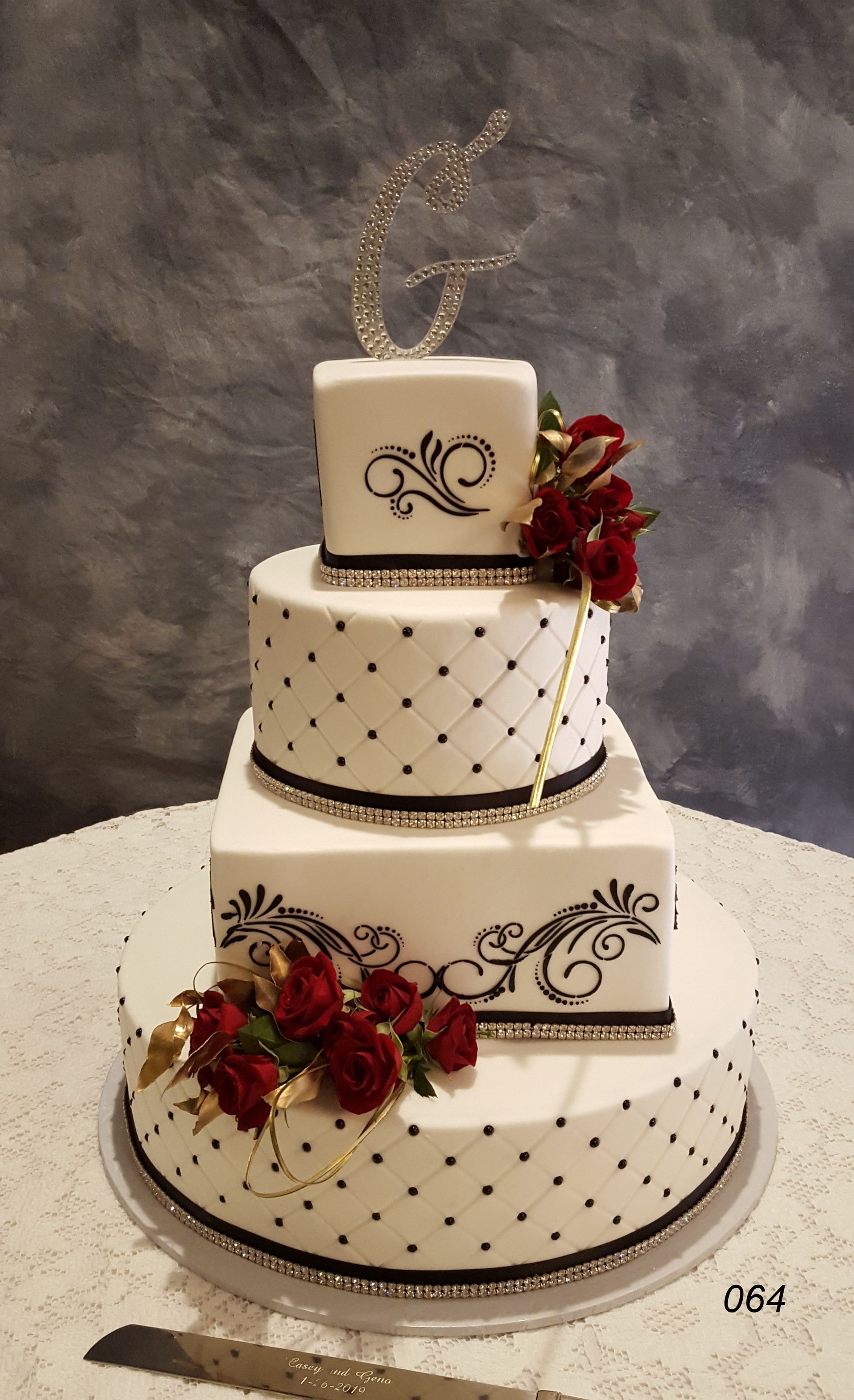 Four-tier white wedding cake with black scroll designs and red floral accents on a stand