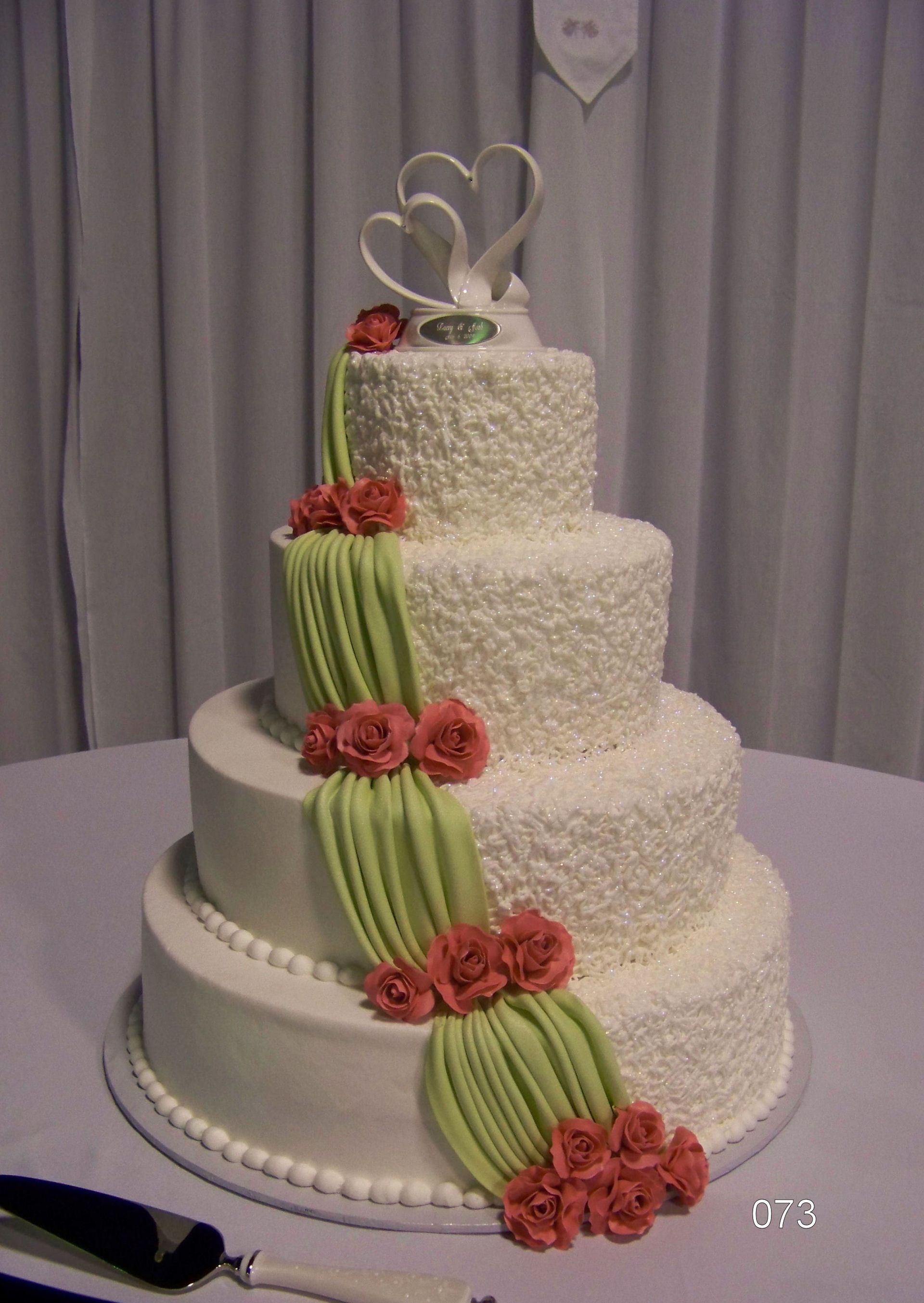 White tiered wedding cake with green ribbon drape, red roses, and silver heart topper