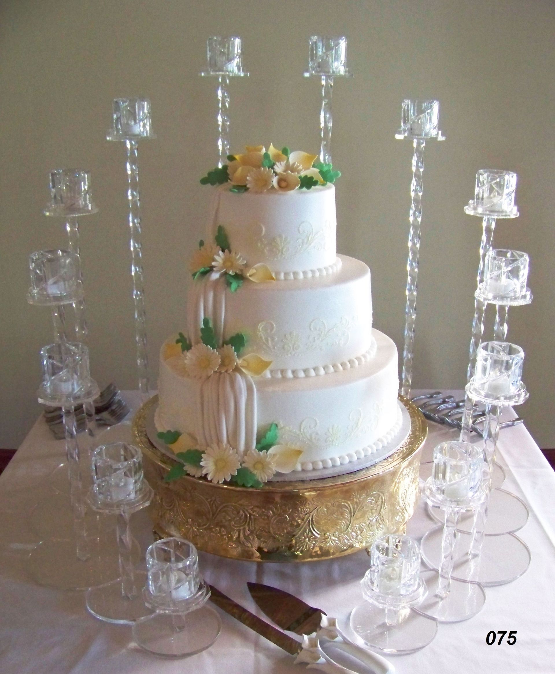 Three-tier white wedding cake with gold accents and flowers, surrounded by stacked glass candle holders