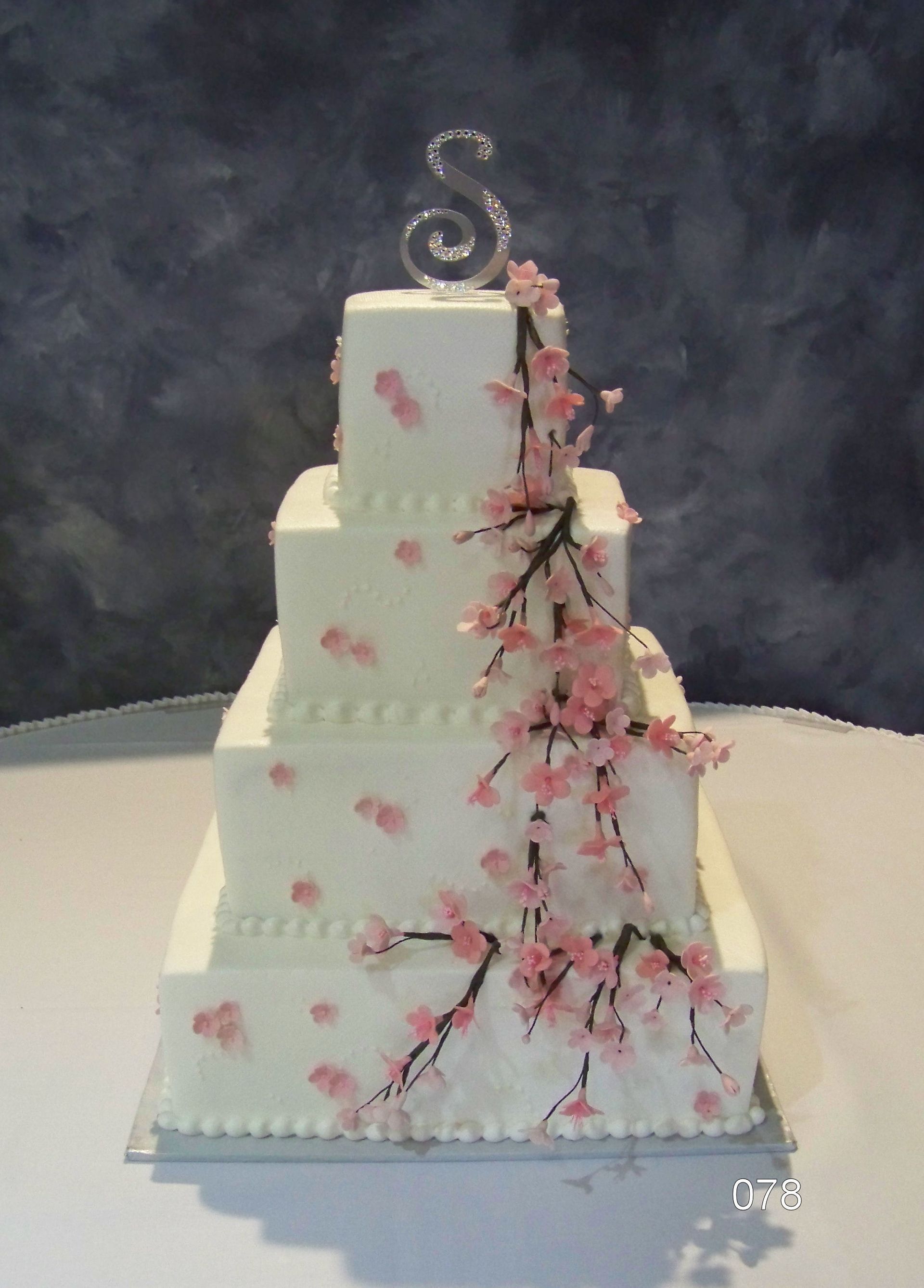 White tiered cake with pink blossoms and a silver swirl topper on a table, against a dark backdrop
