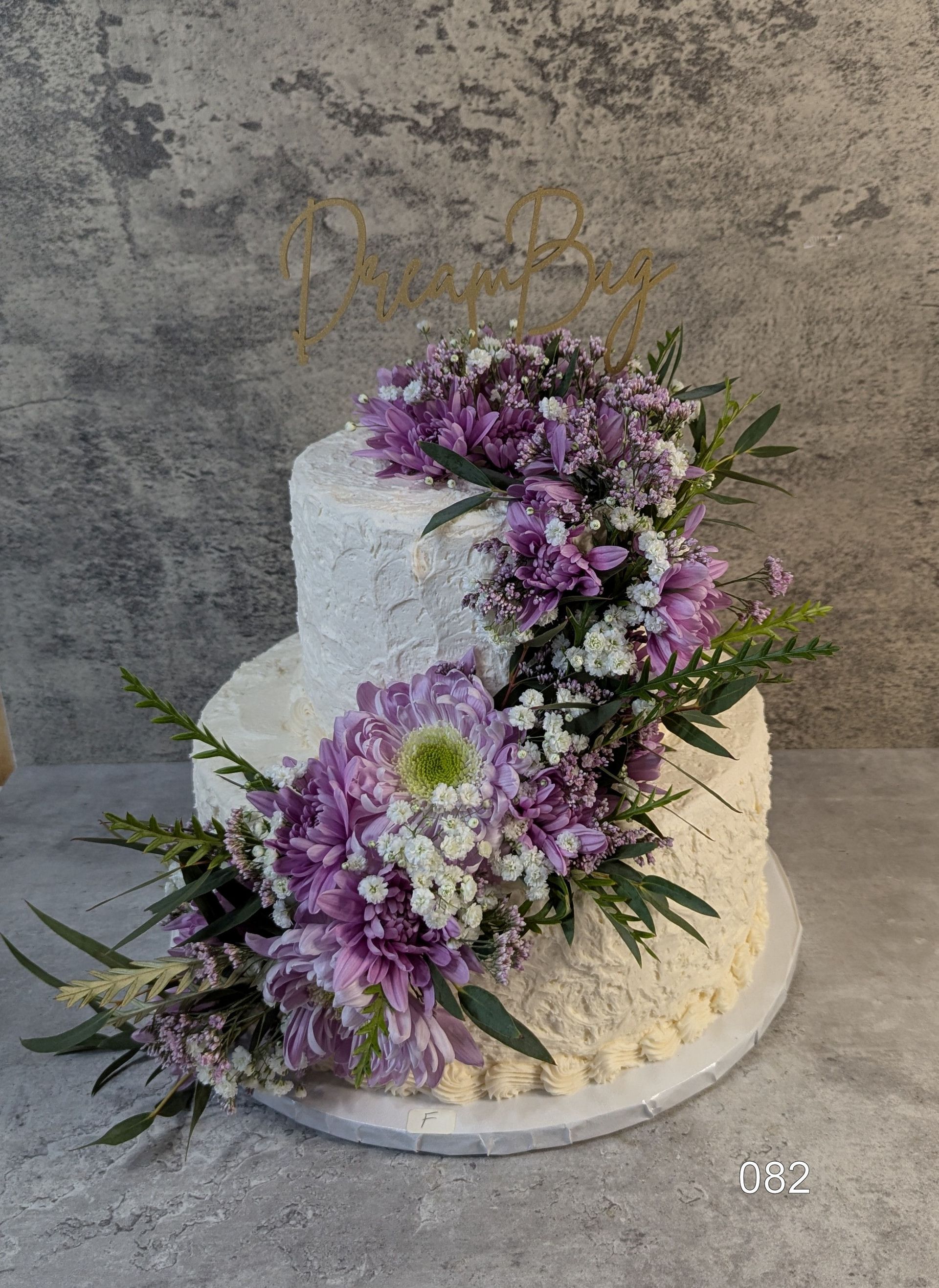 Two-tier white cake with purple and white flowers and greenery on a round stand