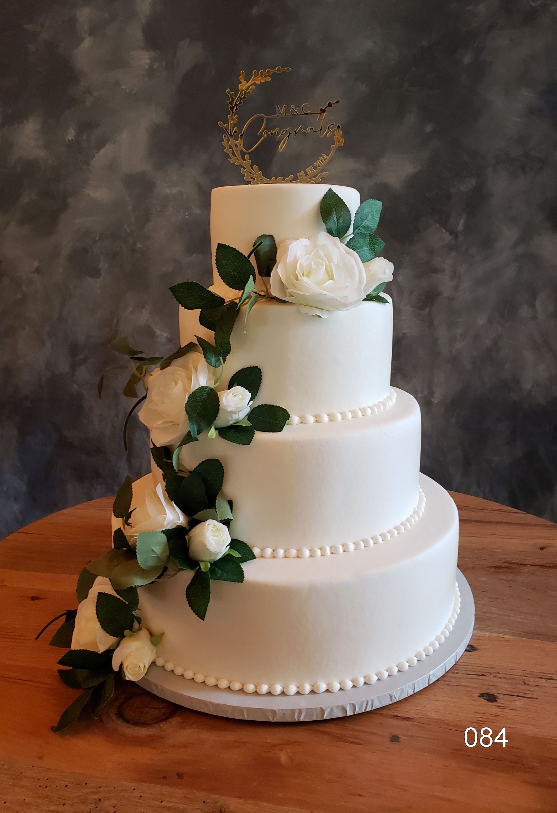 White four-tier wedding cake with white roses, greenery, and gold topper on a wooden table