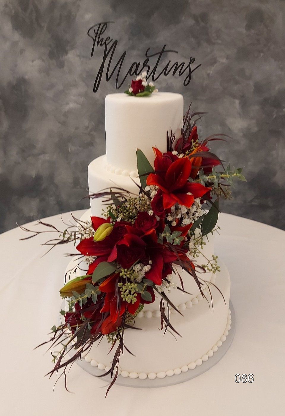 White wedding cake with cascading red floral arrangement and gold accents on a gray backdrop