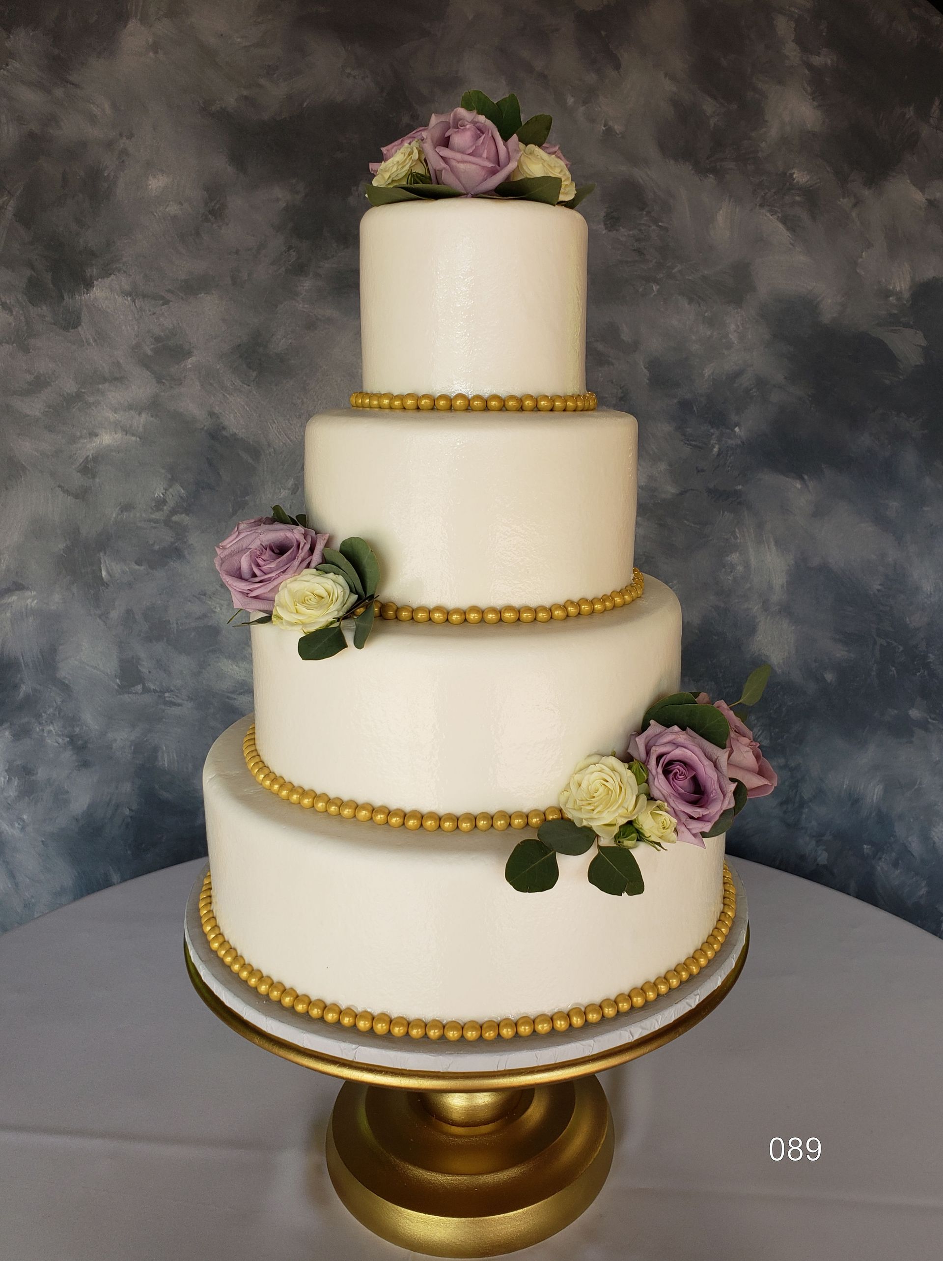 Four-tier white wedding cake with gold beaded trim and purple flowers on a gold stand.
