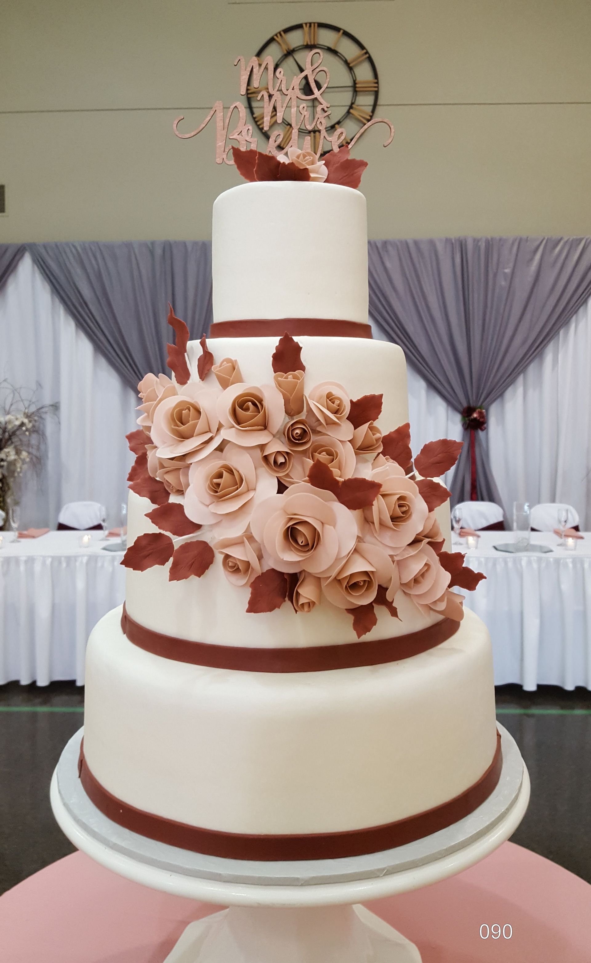 Three-tier white wedding cake with pink flowers and a round topper on a table with draped curtains.