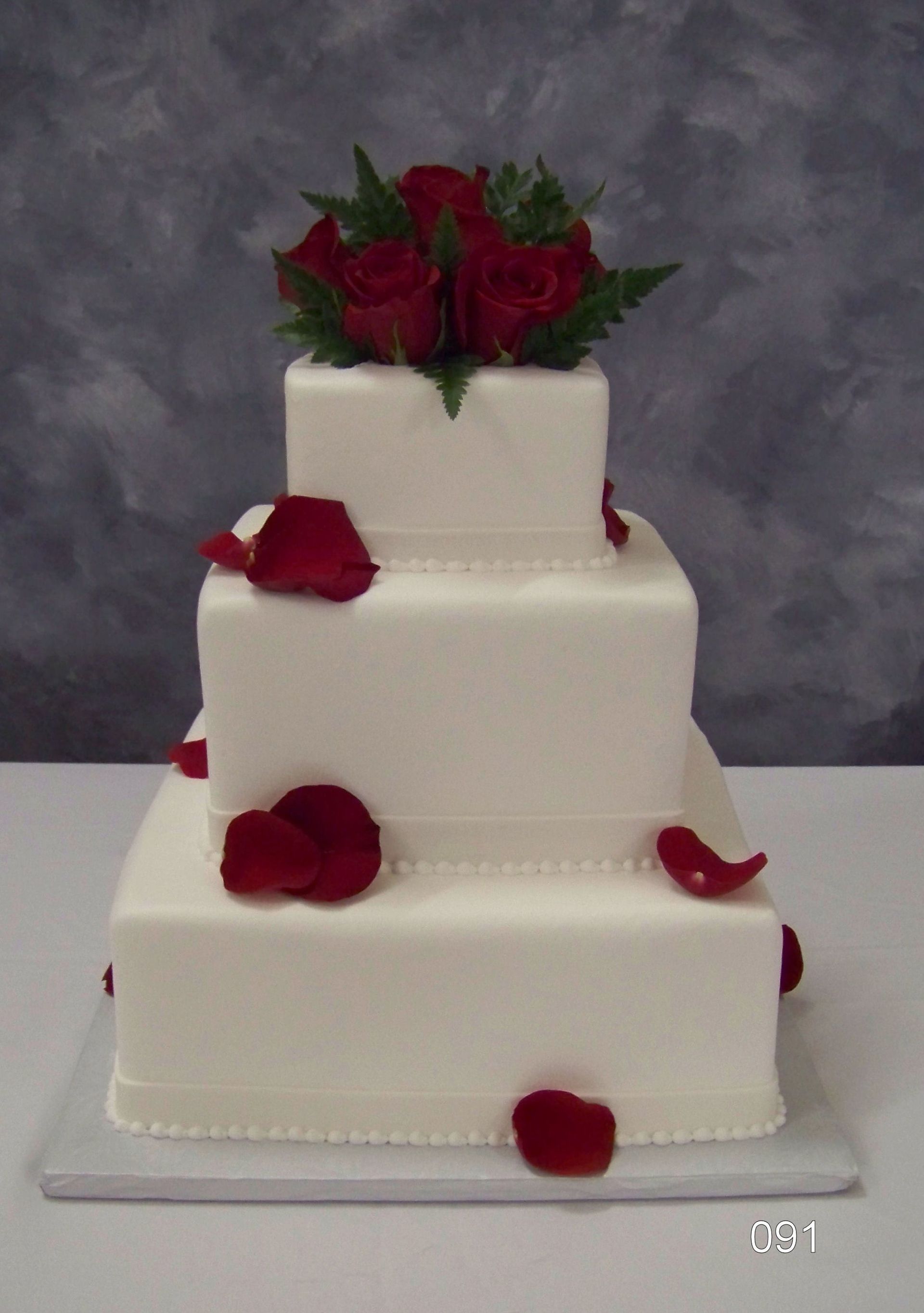 Three-tier white wedding cake with red rose petals and bouquet topper on gray backdrop