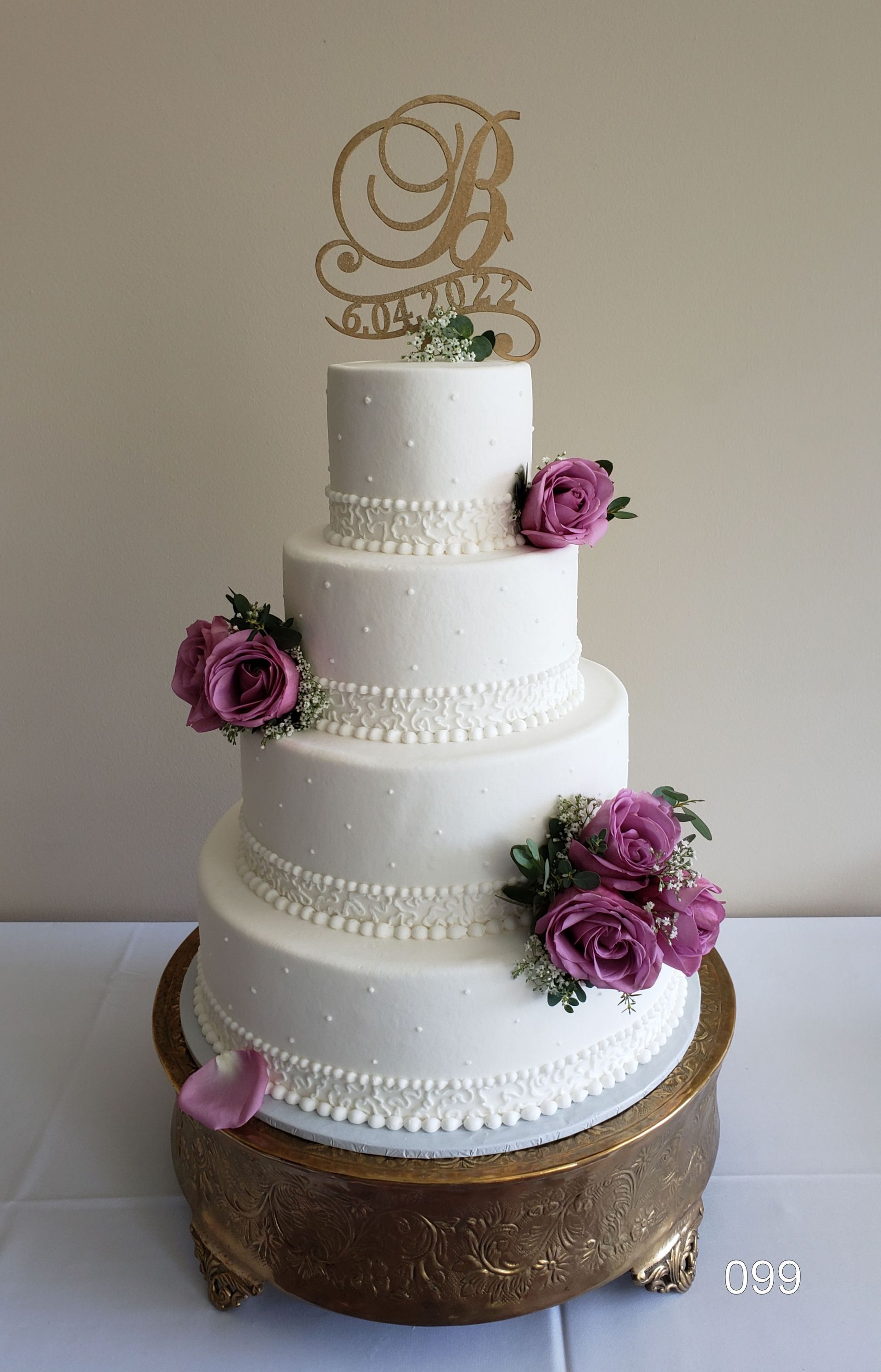 Four-tier white wedding cake with purple roses and gold monogram topper on a rustic base