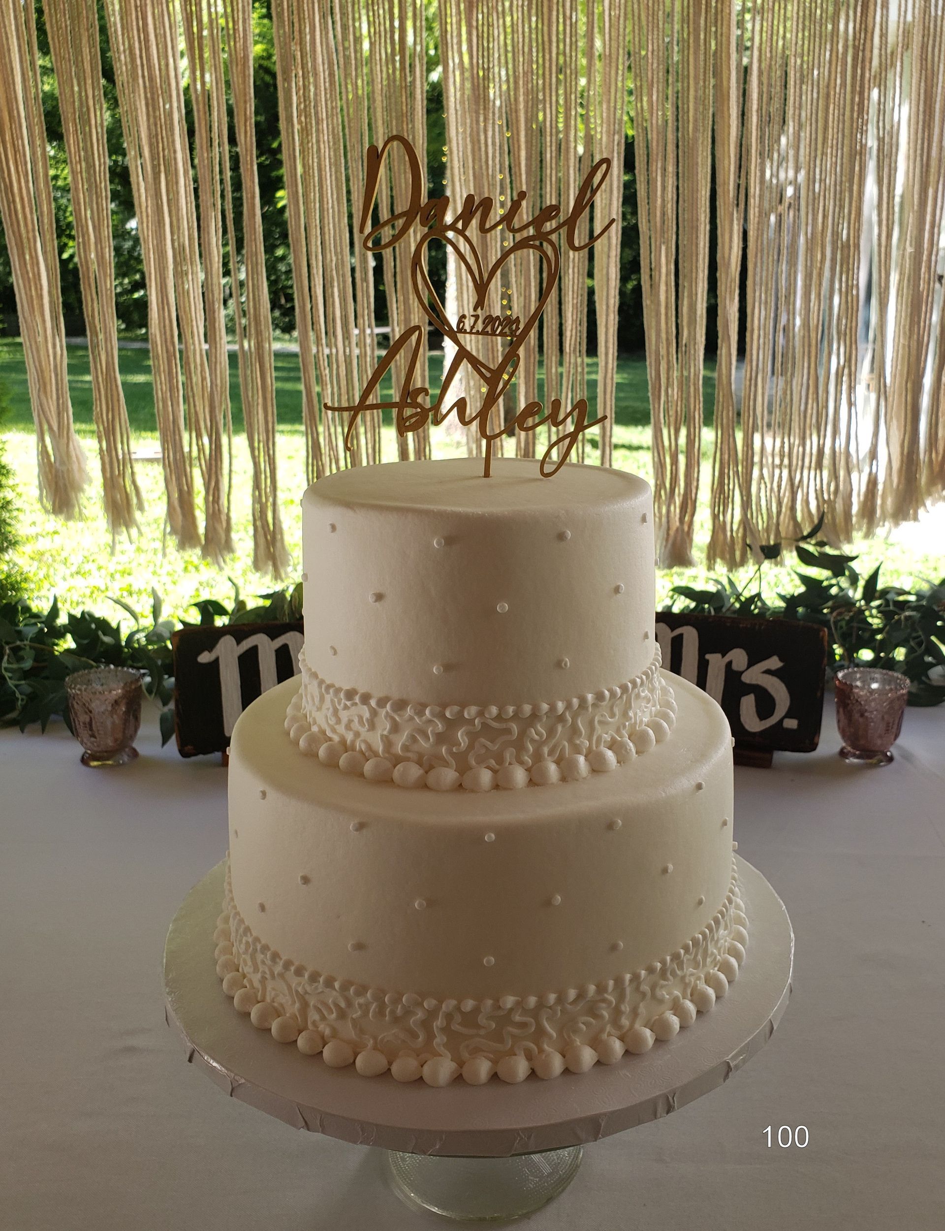 Two-tier white wedding cake with pearl trim and bronze topper on a decorated table outdoors
