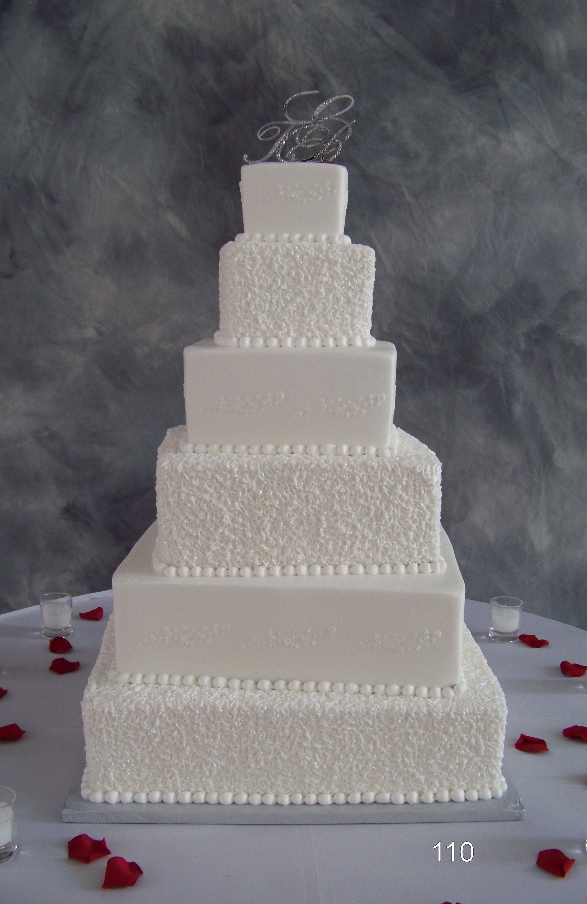 White tiered wedding cake on a table, decorated with pearls and red rose petals against a smoky gray backdrop
