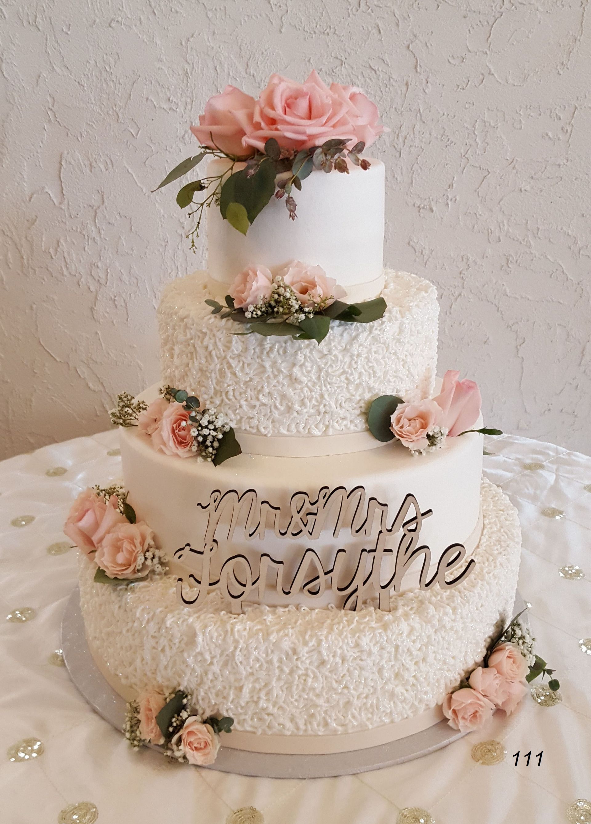 Three-tier white floral cake with pink roses and gold “all the little” topper on a white table.