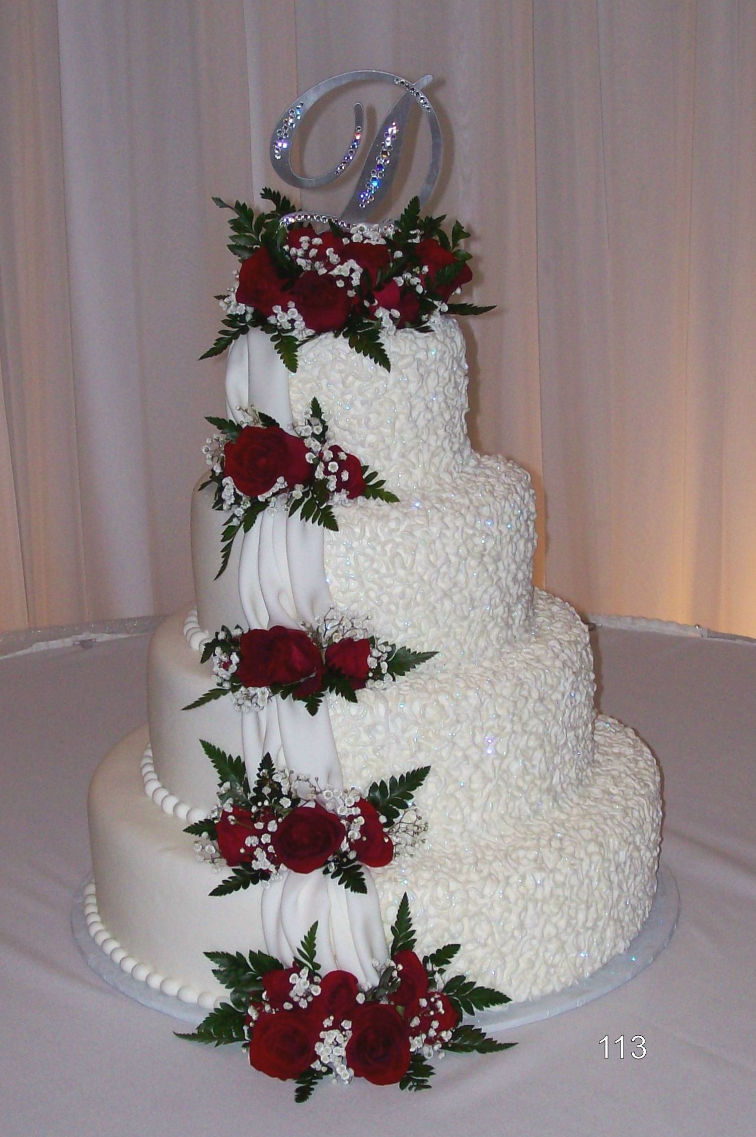 Four-tier white wedding cake with red rose decorations and a silver monogram topper