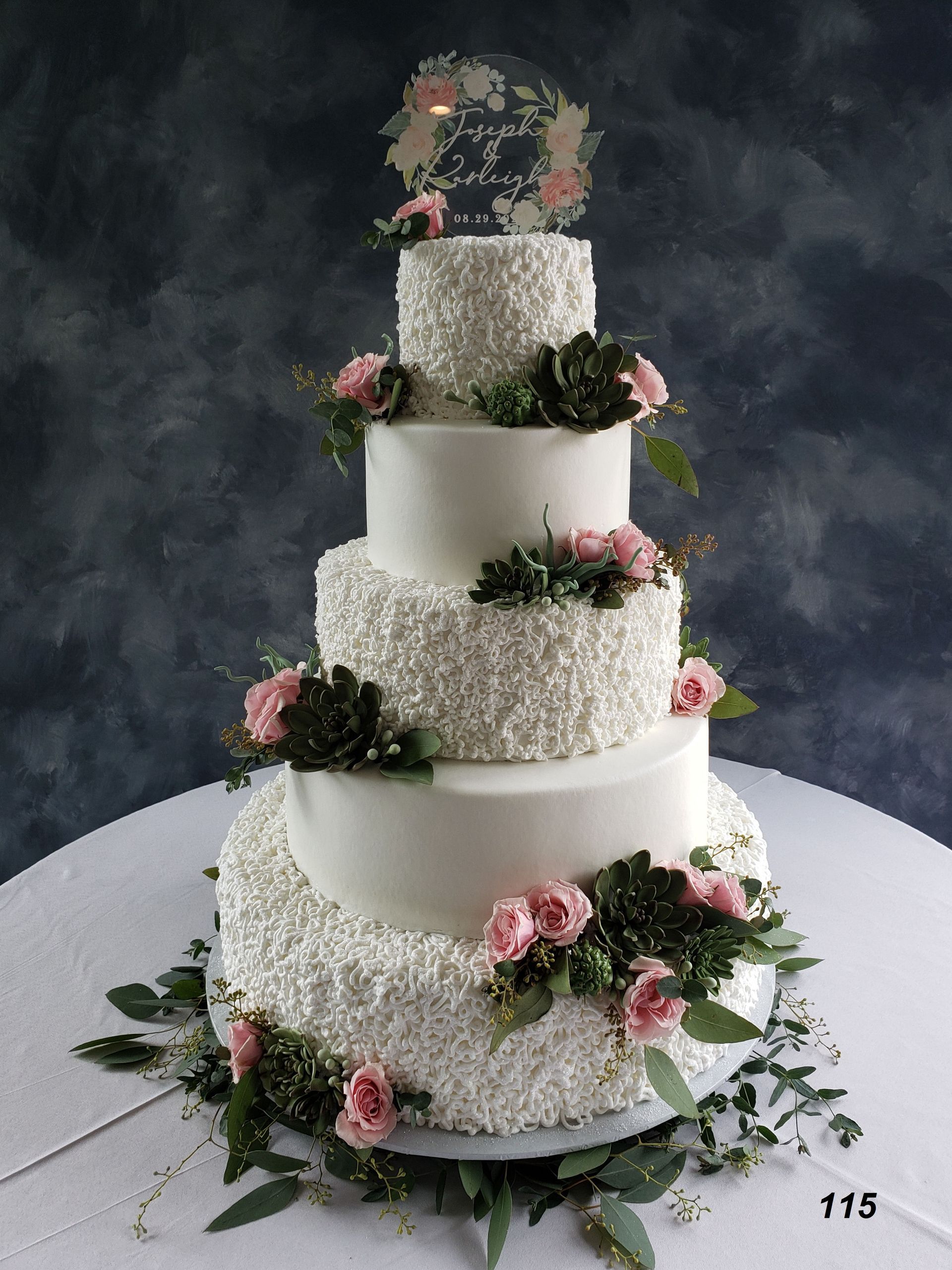 Tall tiered white floral wedding cake with pink roses on a white table against a dark backdrop