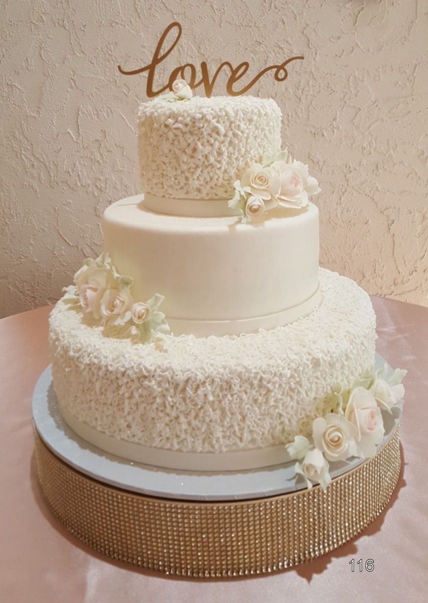 Three-tier white wedding cake with white flowers and a gold “love” topper on a sparkling base
