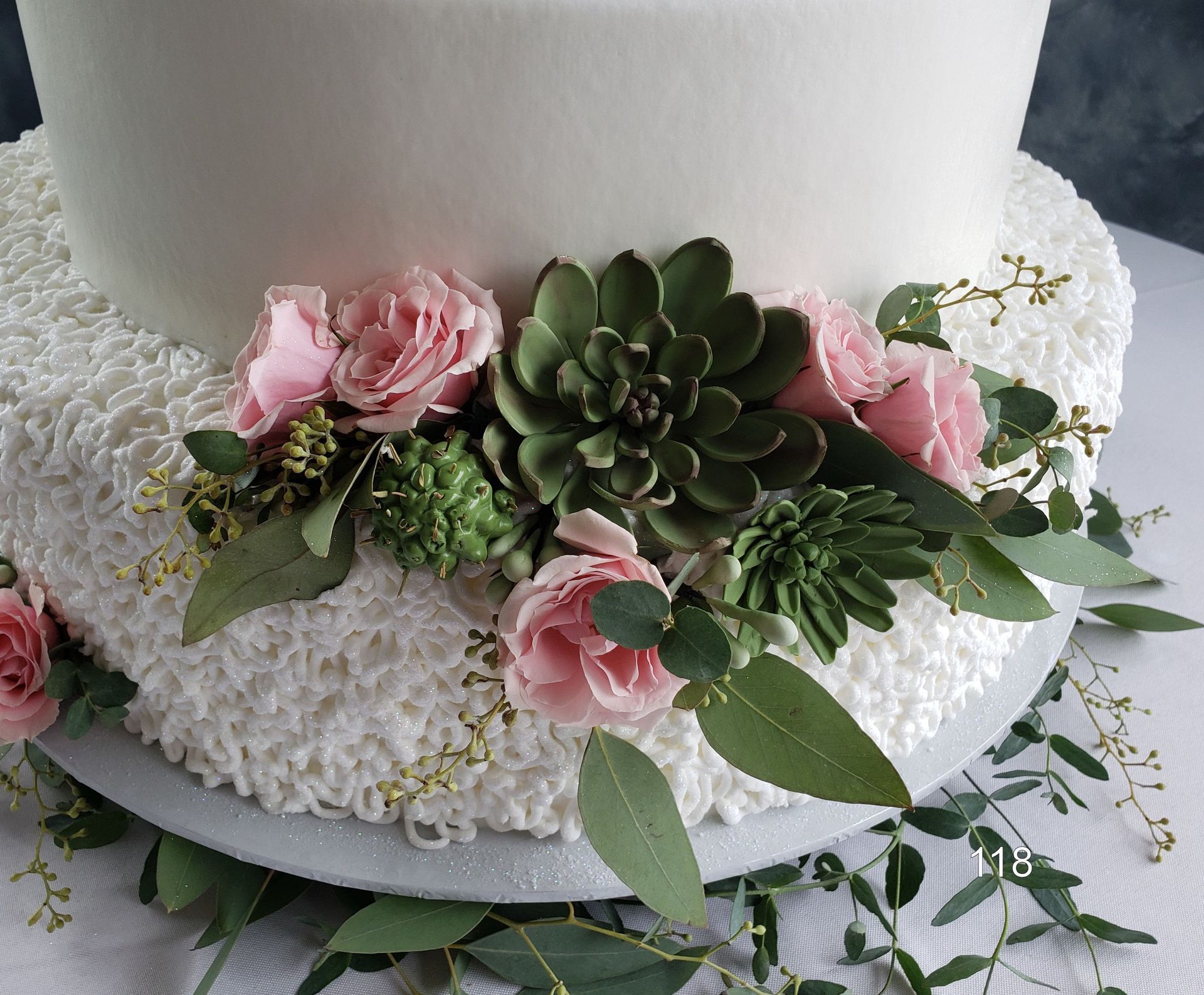 White cake decorated with pink roses, green succulents, and white flowers on a textured white base