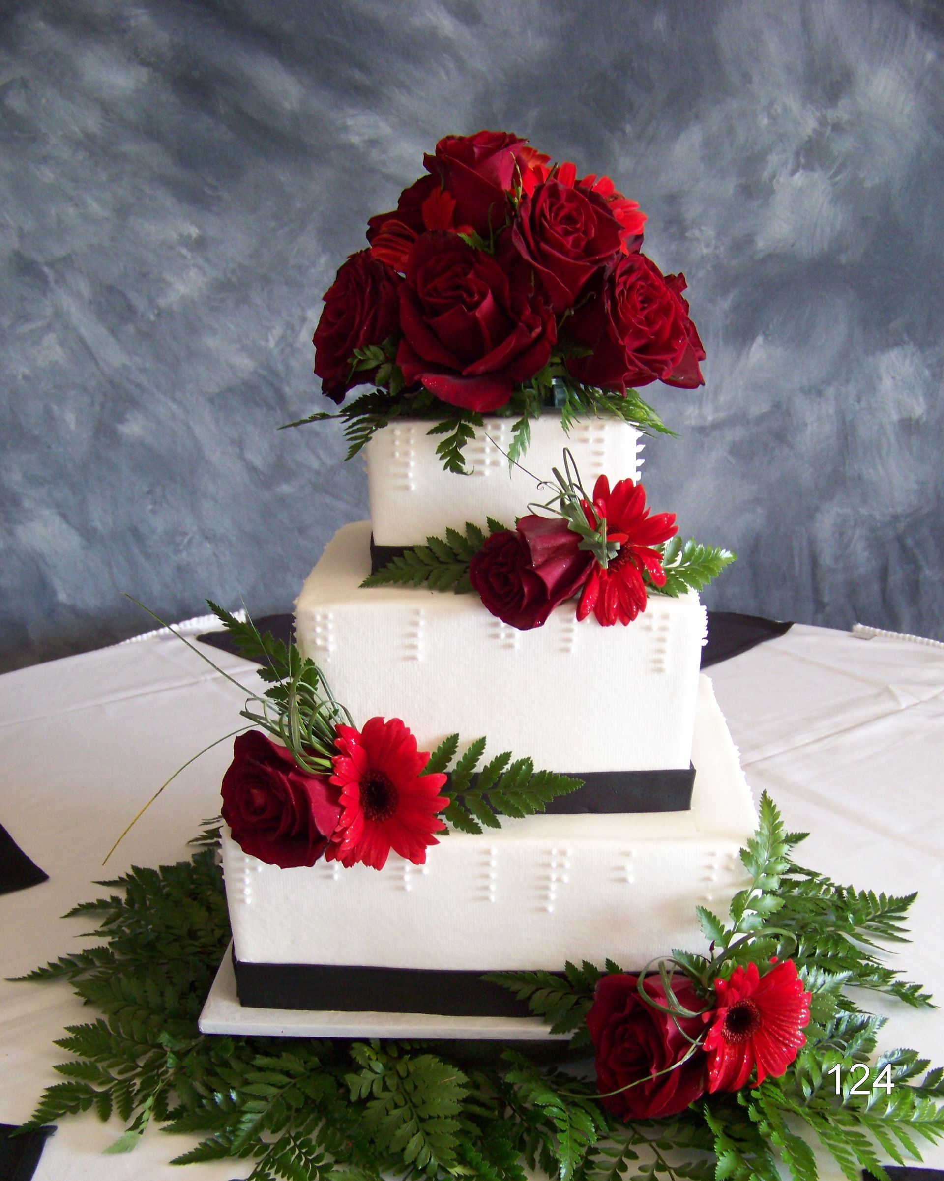 Three-tier white cake with red roses and black ribbon accents on a white tablecloth.