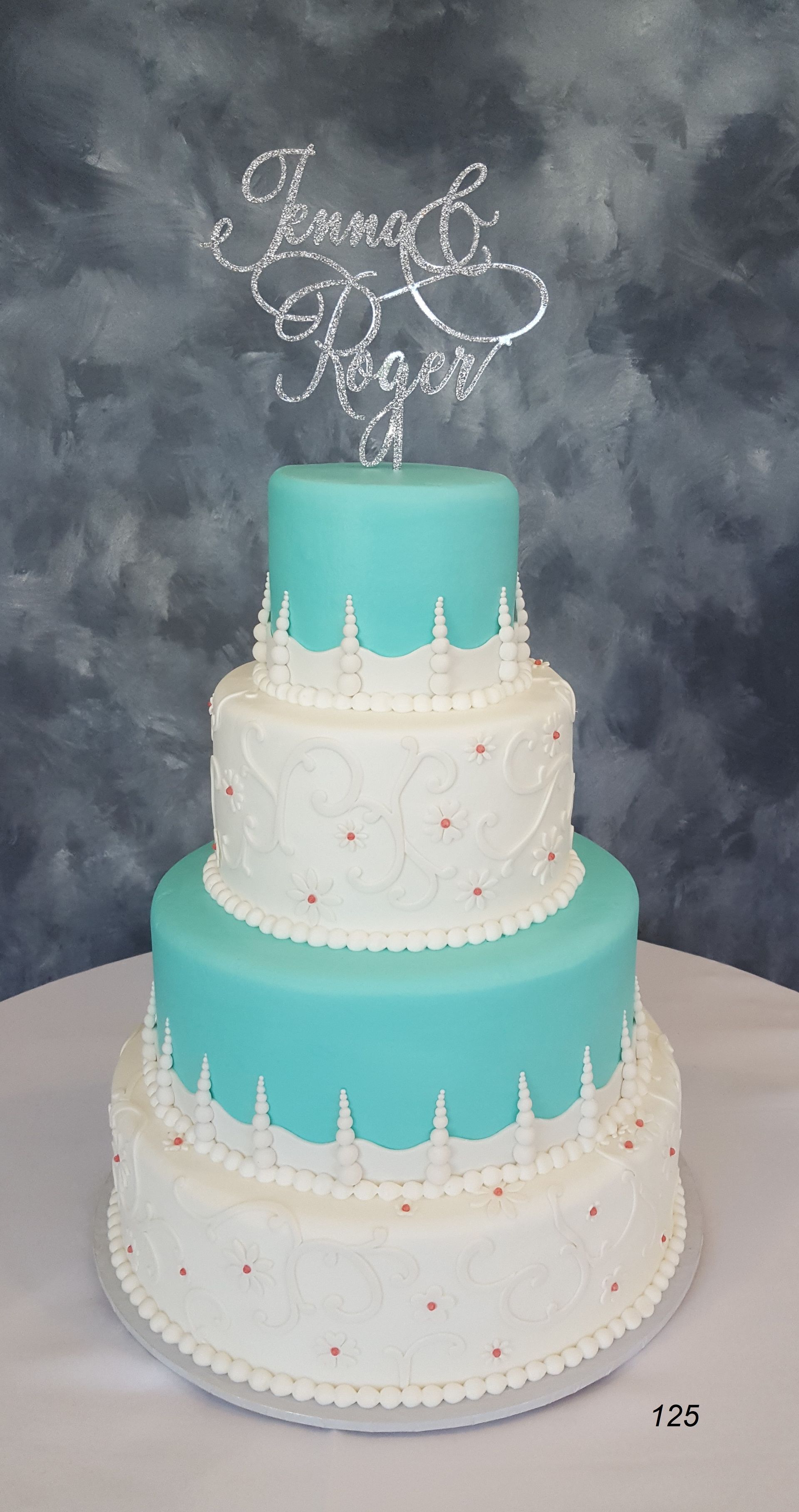 Tiered white and turquoise wedding cake with a “Happy 50th” topper on a gray backdrop