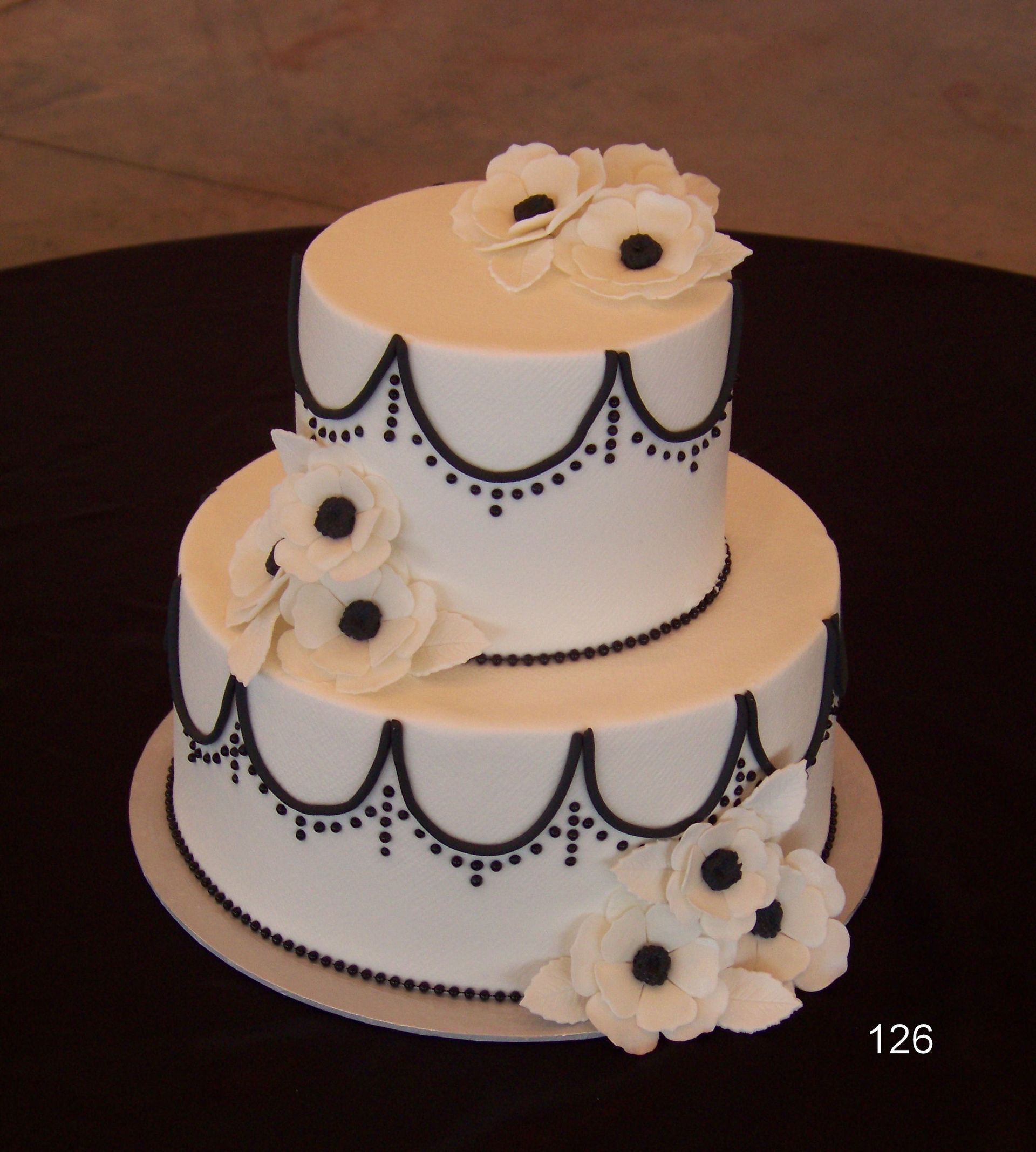 Two-tier white wedding cake with black dotted trim and white flowers on a dark table