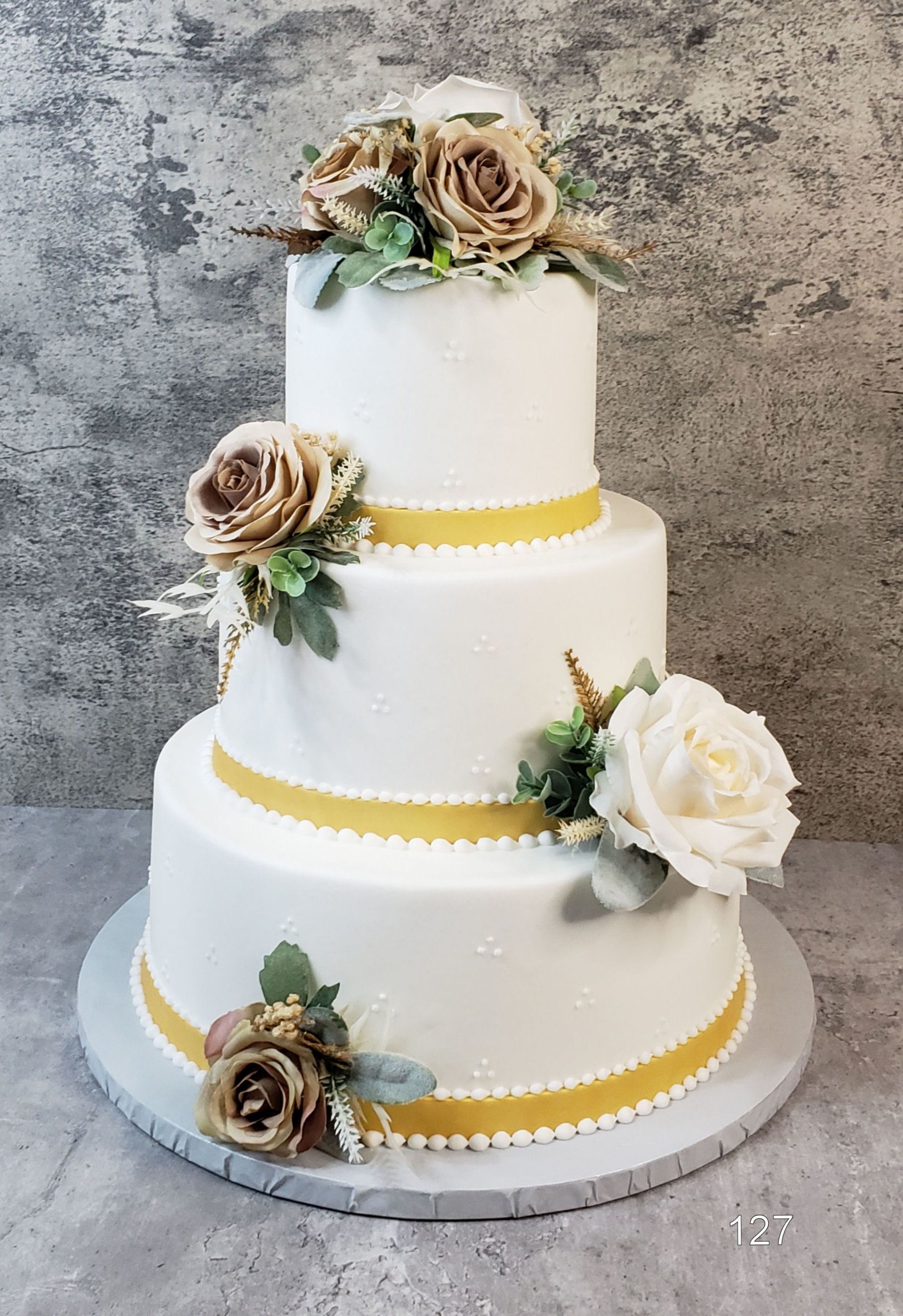 Three-tier white cake with yellow ribbon trim and beige and white flowers on a gray cake board