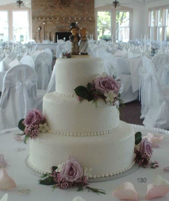 White tiered wedding cake with purple flowers and a topper on a decorated reception table