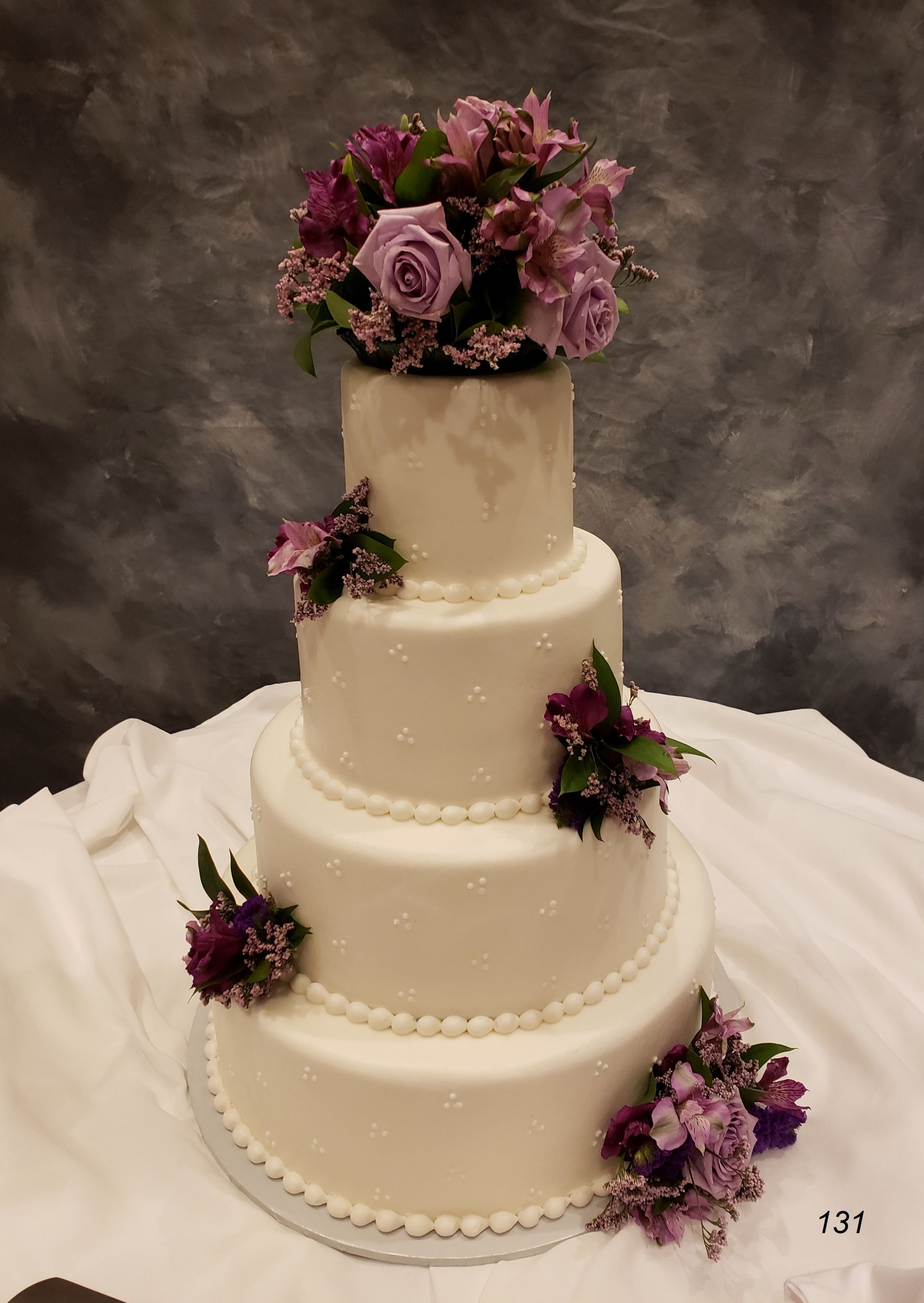 Four-tier ivory wedding cake with purple floral decorations on a white draped table