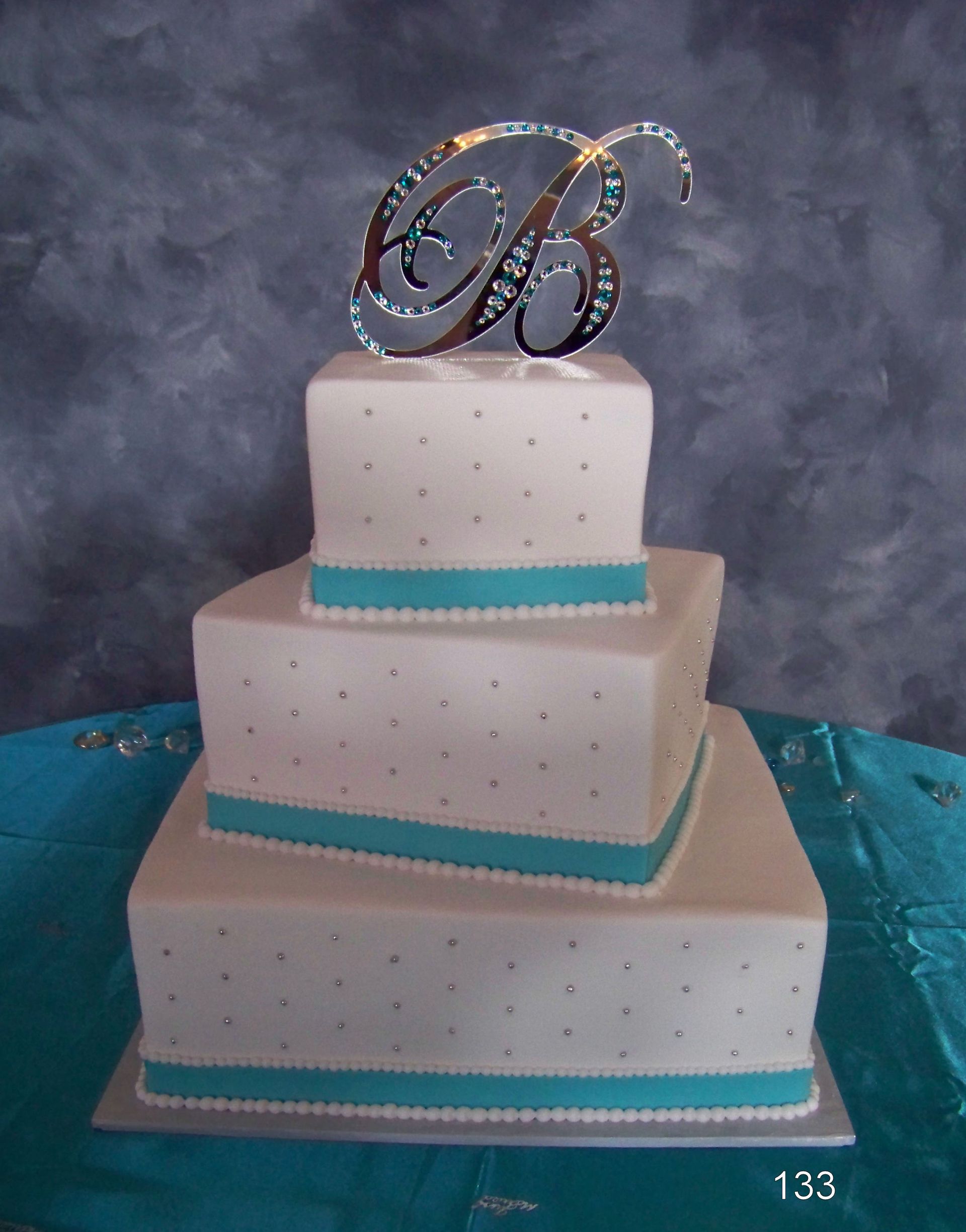 Three-tier white wedding cake with turquoise trim and a sparkling monogram topper on a blue tablecloth