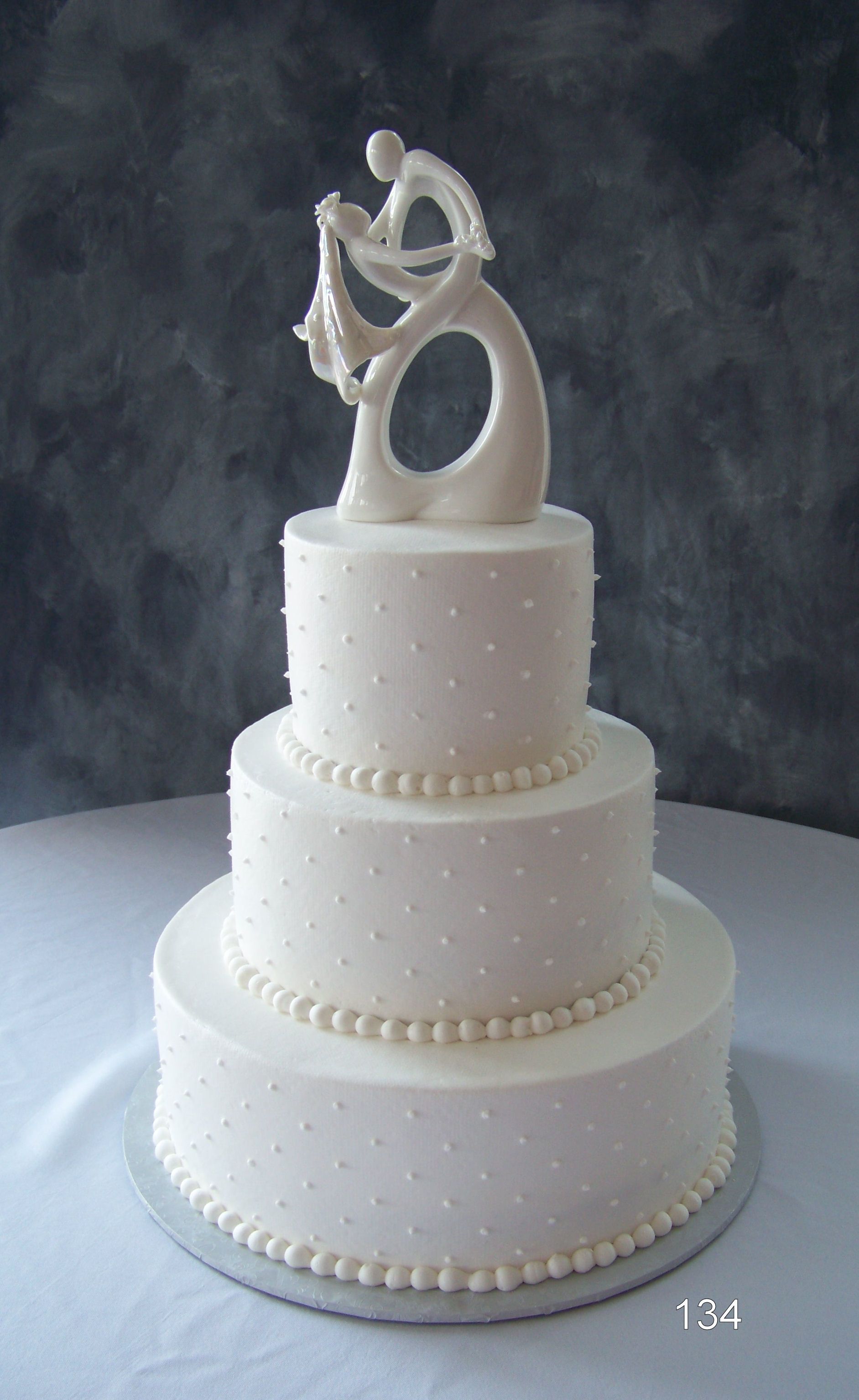 Three-tier white wedding cake with pearl trim and abstract silver topper on a dark background