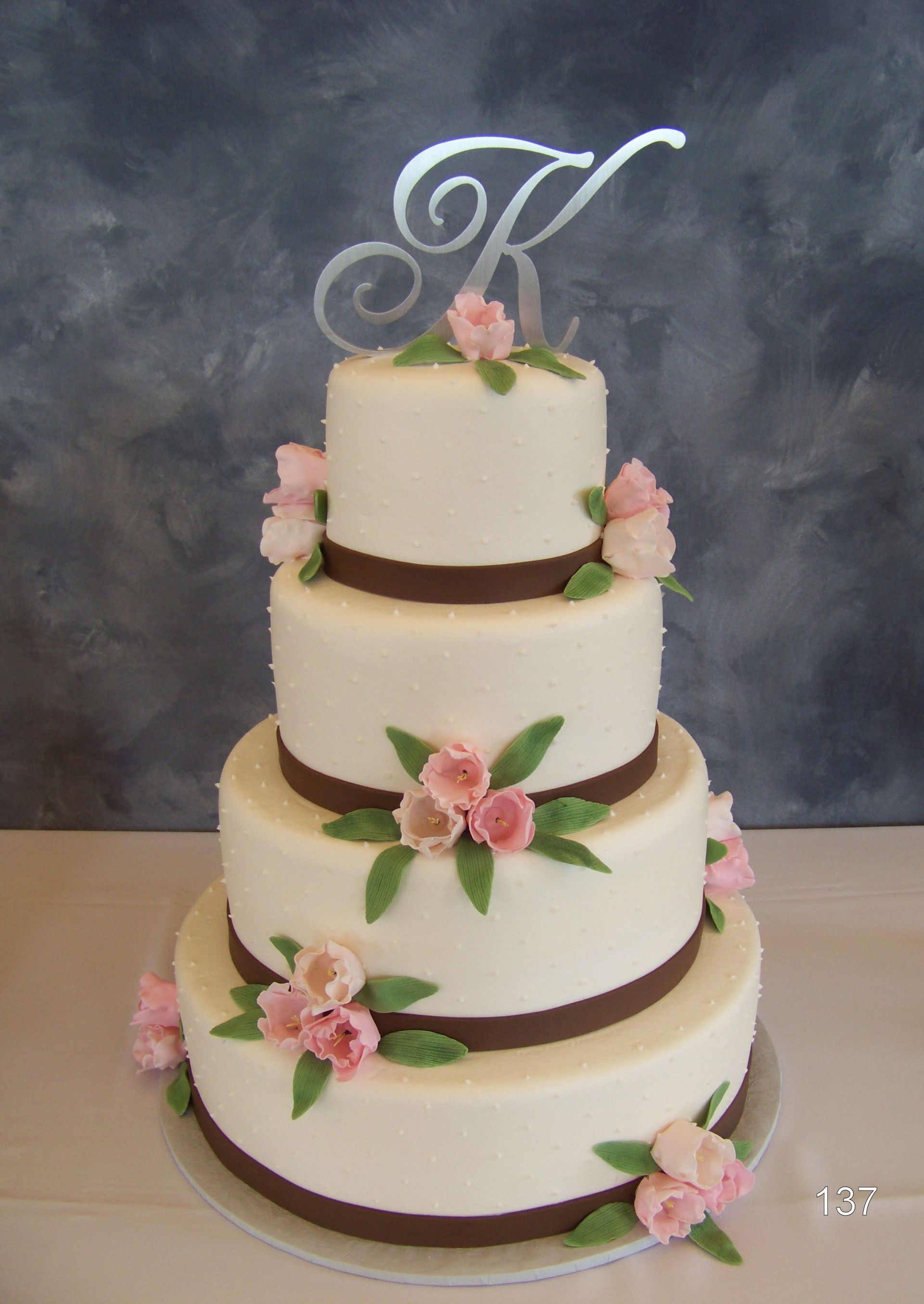 Four-tier white wedding cake with pink flowers and a silver monogram topper against a gray backdrop
